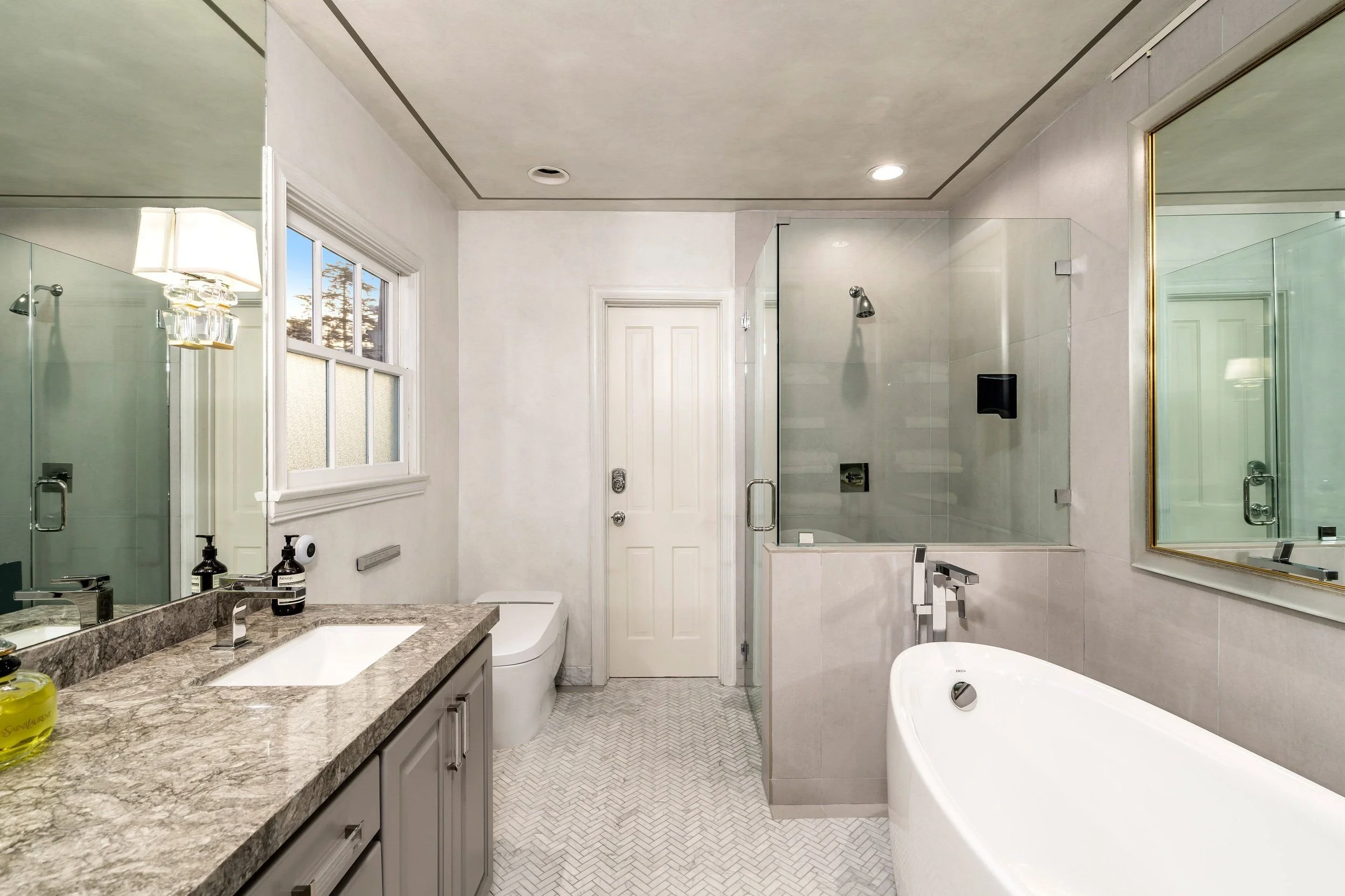 A modern bathroom with a double sink vanity, granite countertop, large mirror, and a bathtub. There is a window with a view of trees, a glass shower enclosure, and white tiled walls and floor.