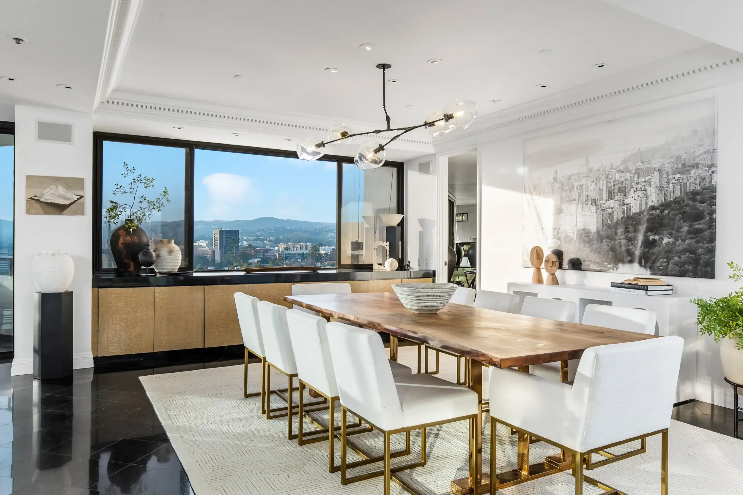 Modern dining room with a large wooden table, white chairs, black tiled floor, and large windows showing a cityscape view. Decor includes vases, sculptures, and art on the walls.