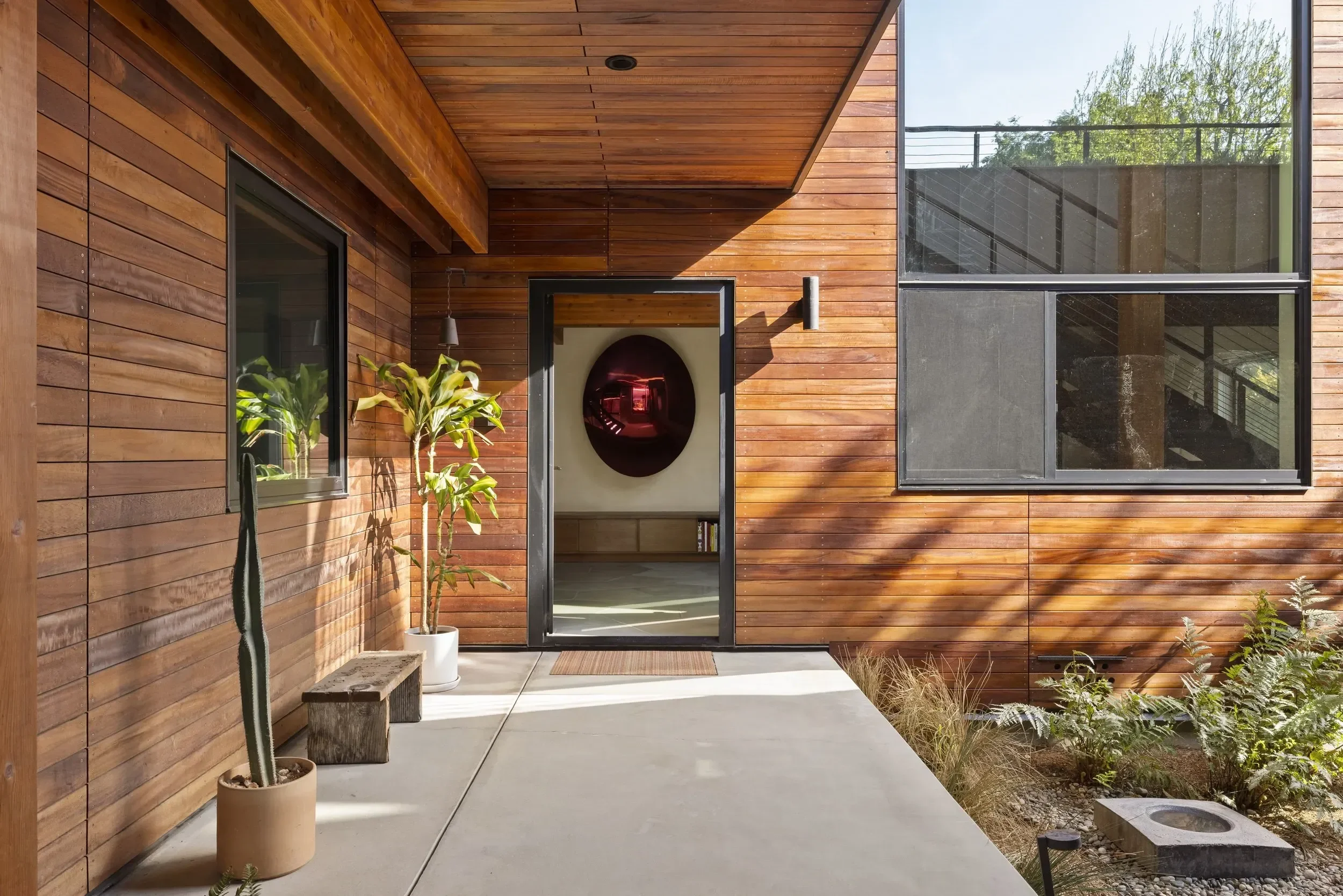 Modern house exterior with wooden siding, large windows, and a small garden area with potted plants and shrubs.
