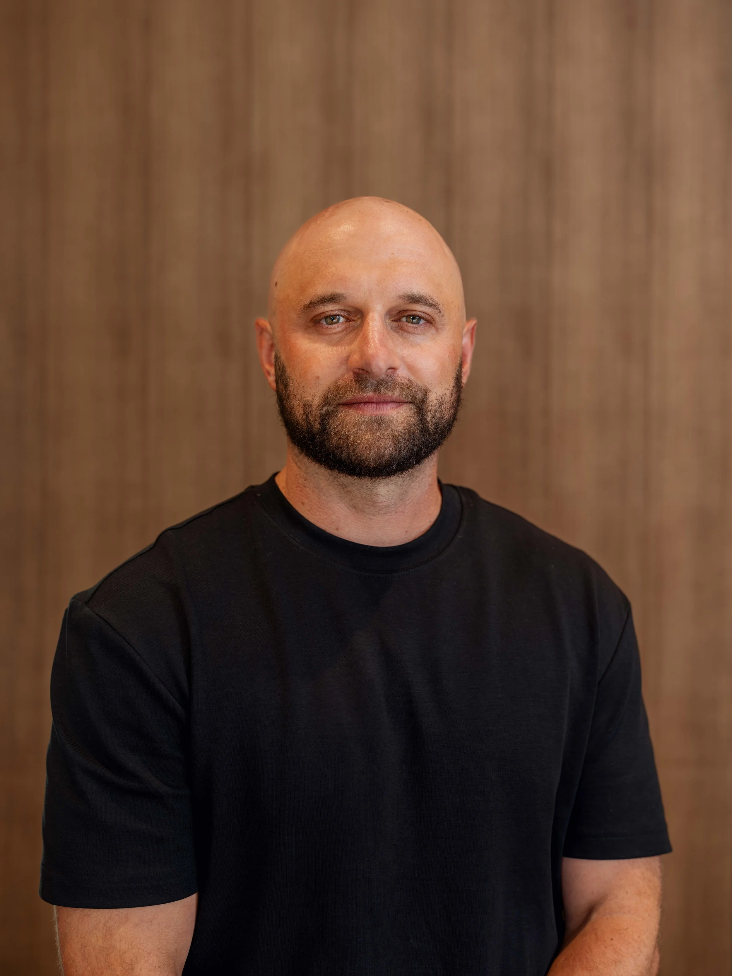 A man with a bald head, beard, and light-colored eyes, wearing a black T-shirt, standing in front of a wooden wall.