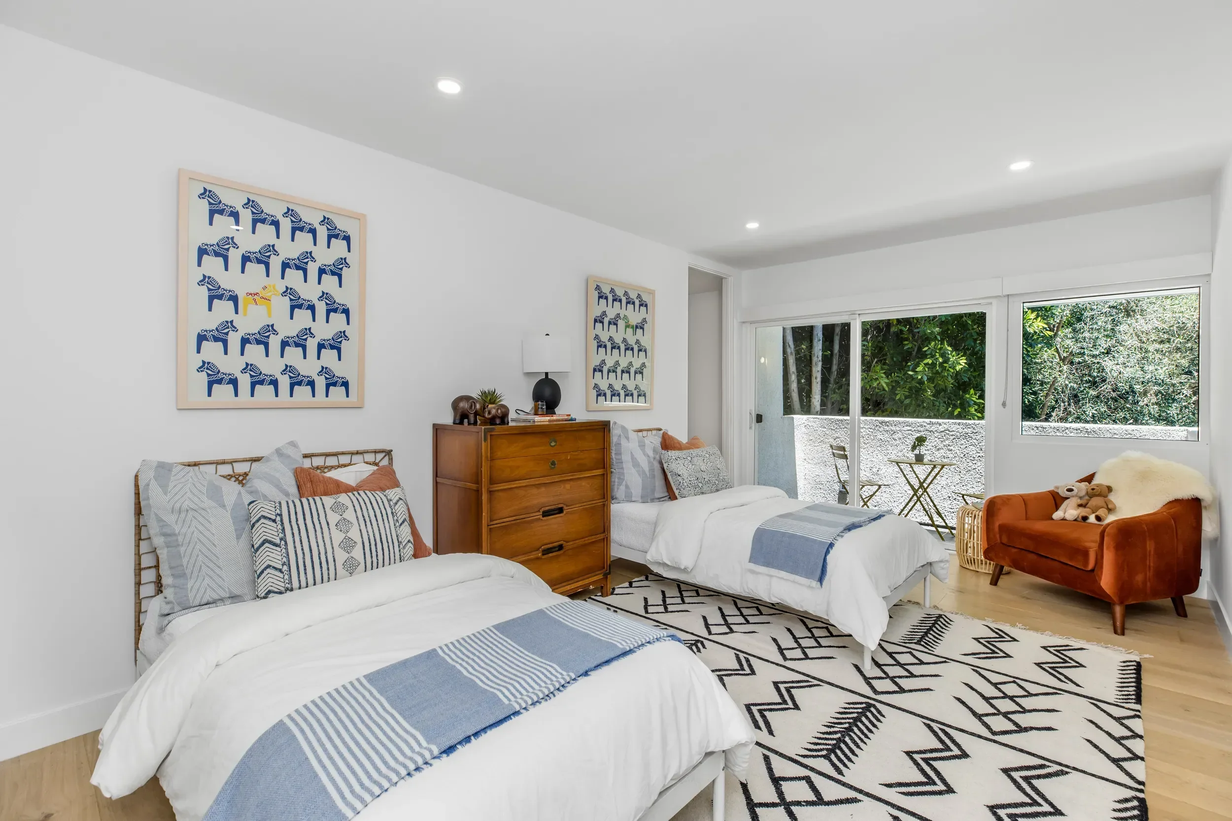 Bright living room with white walls and large window showing outdoor greenery. Three twin beds with white and blue striped bedding and patterned pillows. Wooden dresser with a black lamp, decorative elephant figurines, and books. Wall art featuring b