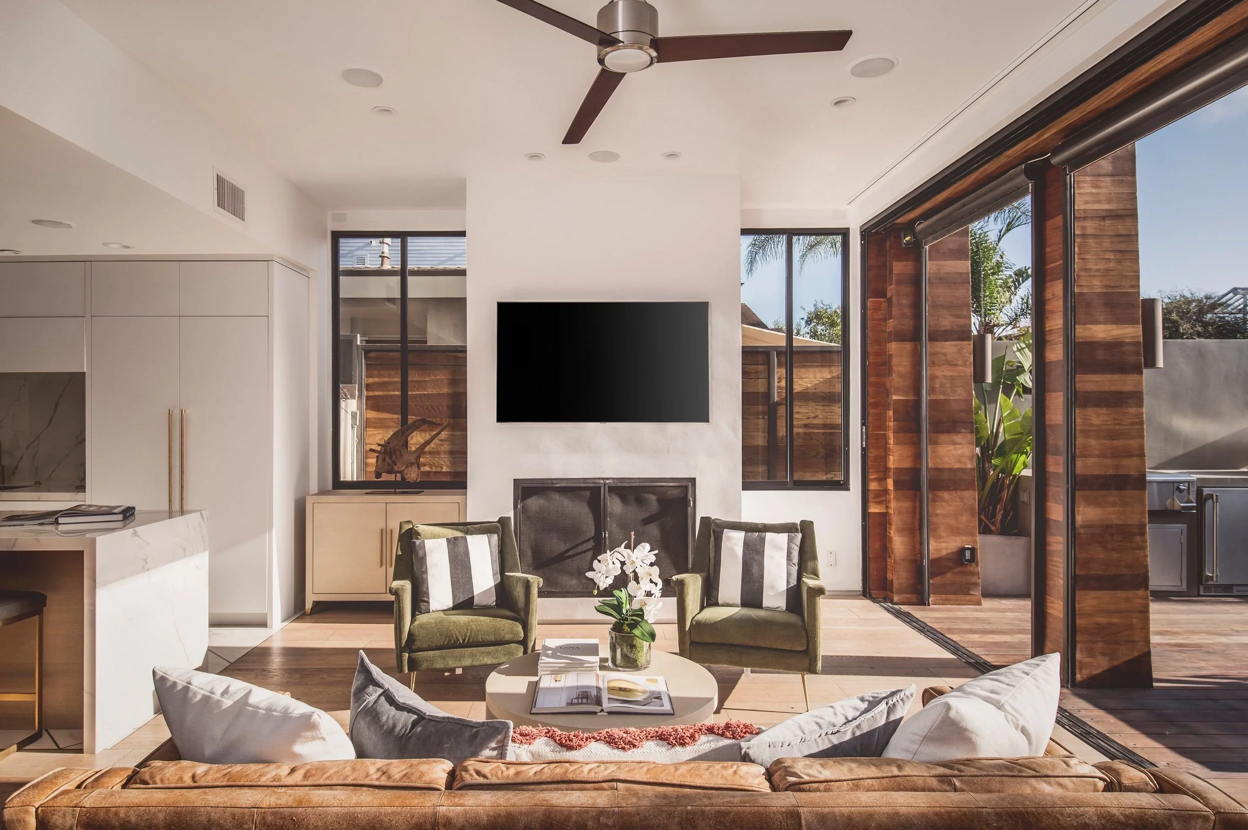 Modern living room with a white fireplace, flat-screen TV, glass doors leading to an outdoor patio, green and black striped chairs, orchids on a coffee table, and a ceiling fan.