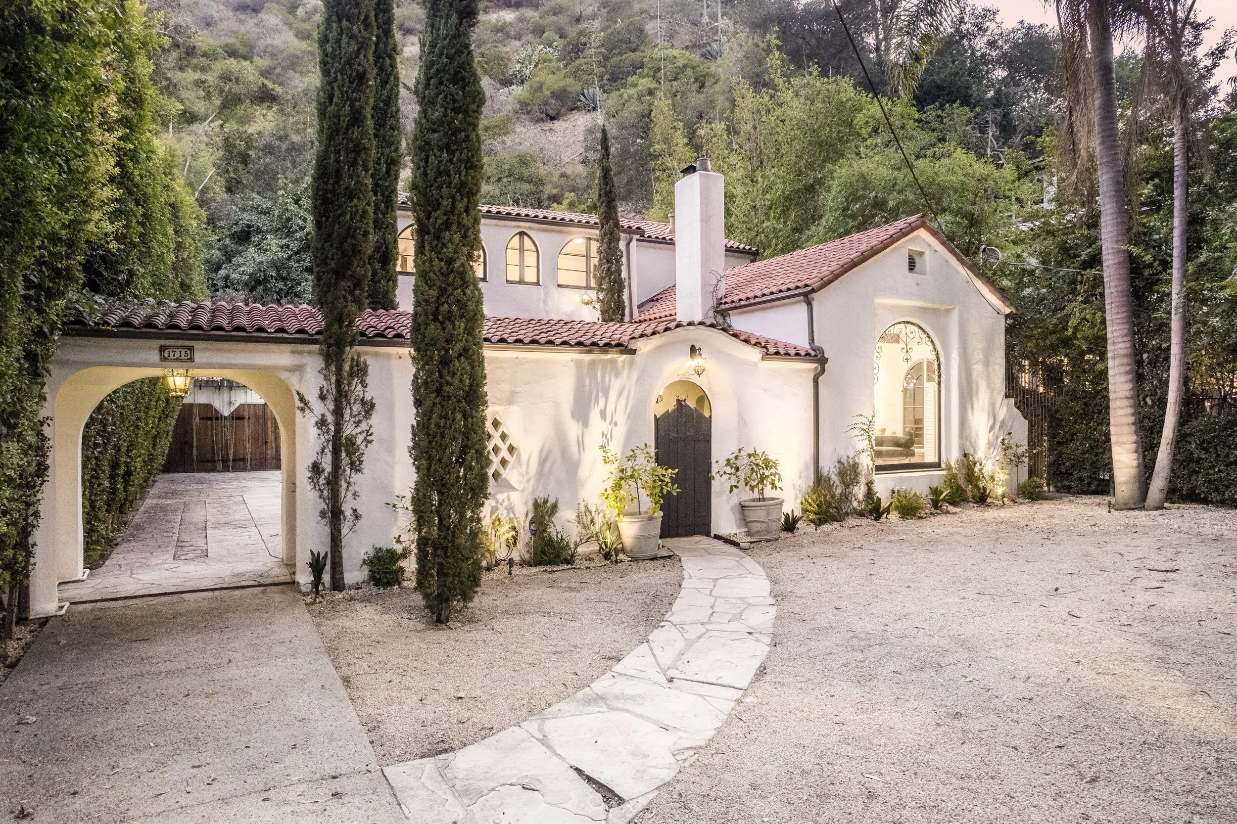 A white house with a red tile roof, arched windows, and a wooden gate, surrounded by trees and plants, with a curved stone pathway leading to the entrance.