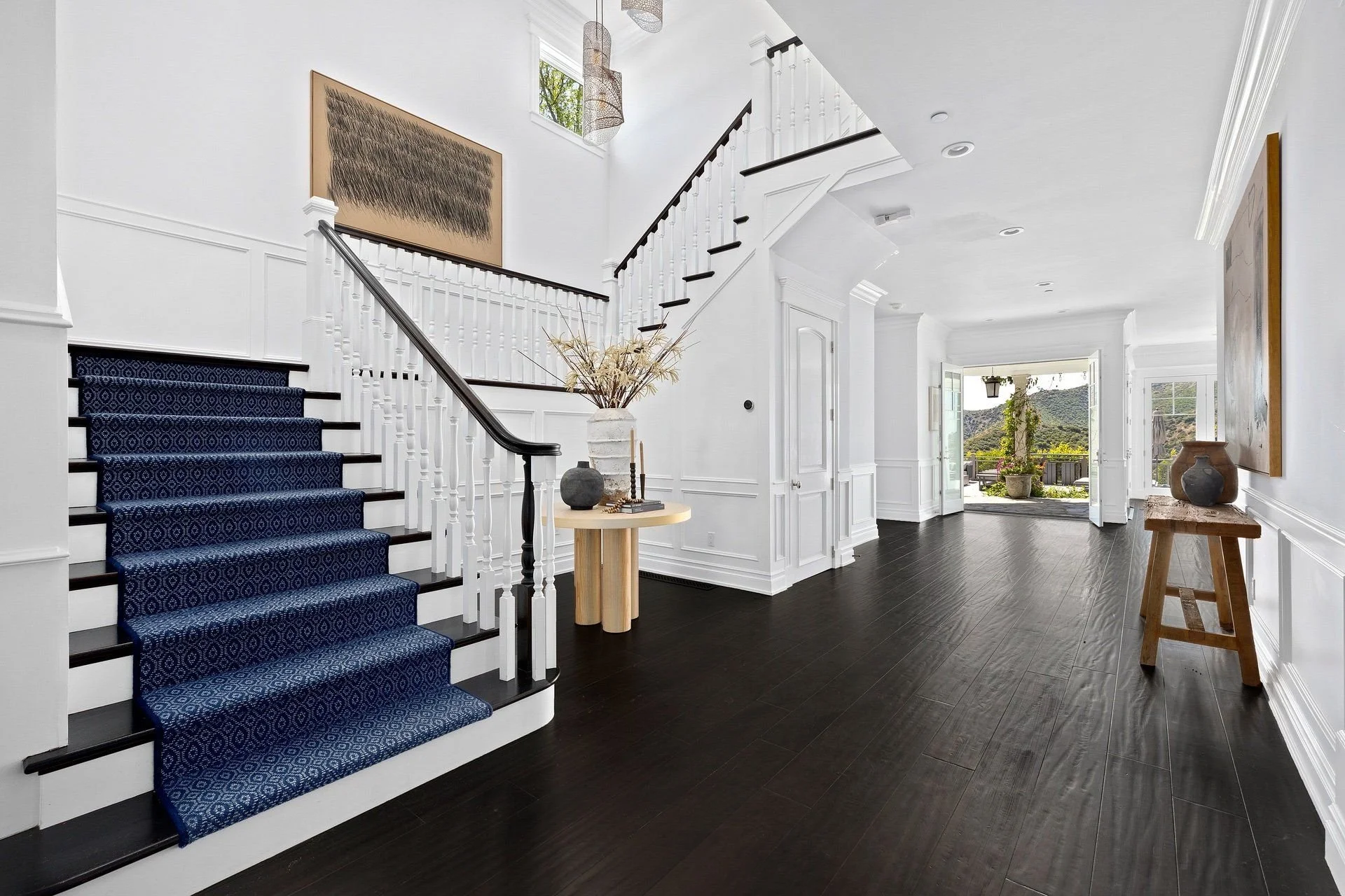 Modern interior entryway with staircase carpeted in blue, white walls, and dark wooden floors. There are decorations including vases, artwork, and a small hallway leading outside to a view of mountains and greenery.