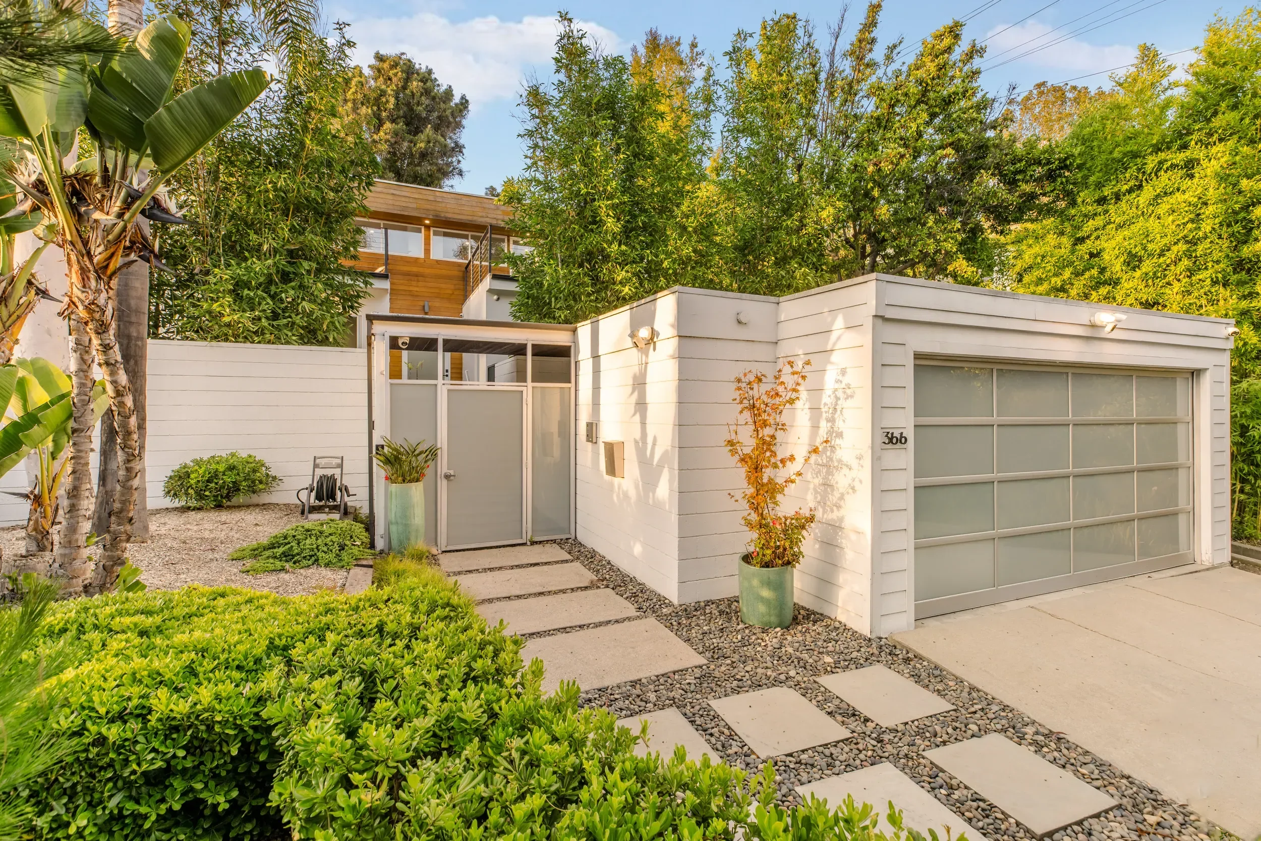 Modern white house with a garage door, surrounded by lush green plants and trees, with a pathway and decorative gravel in the yard.