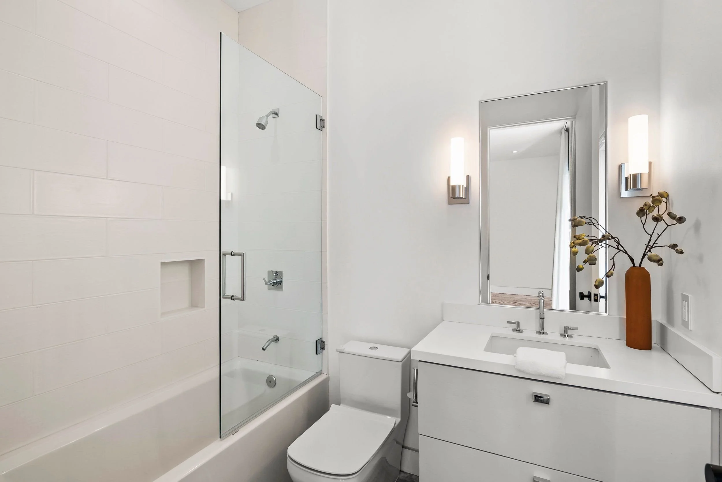Modern bathroom with a white bathtub, a glass shower enclosure, and a sleek rectangular vanity with a large mirror, wall sconces, and a vase with dried decorative branches.
