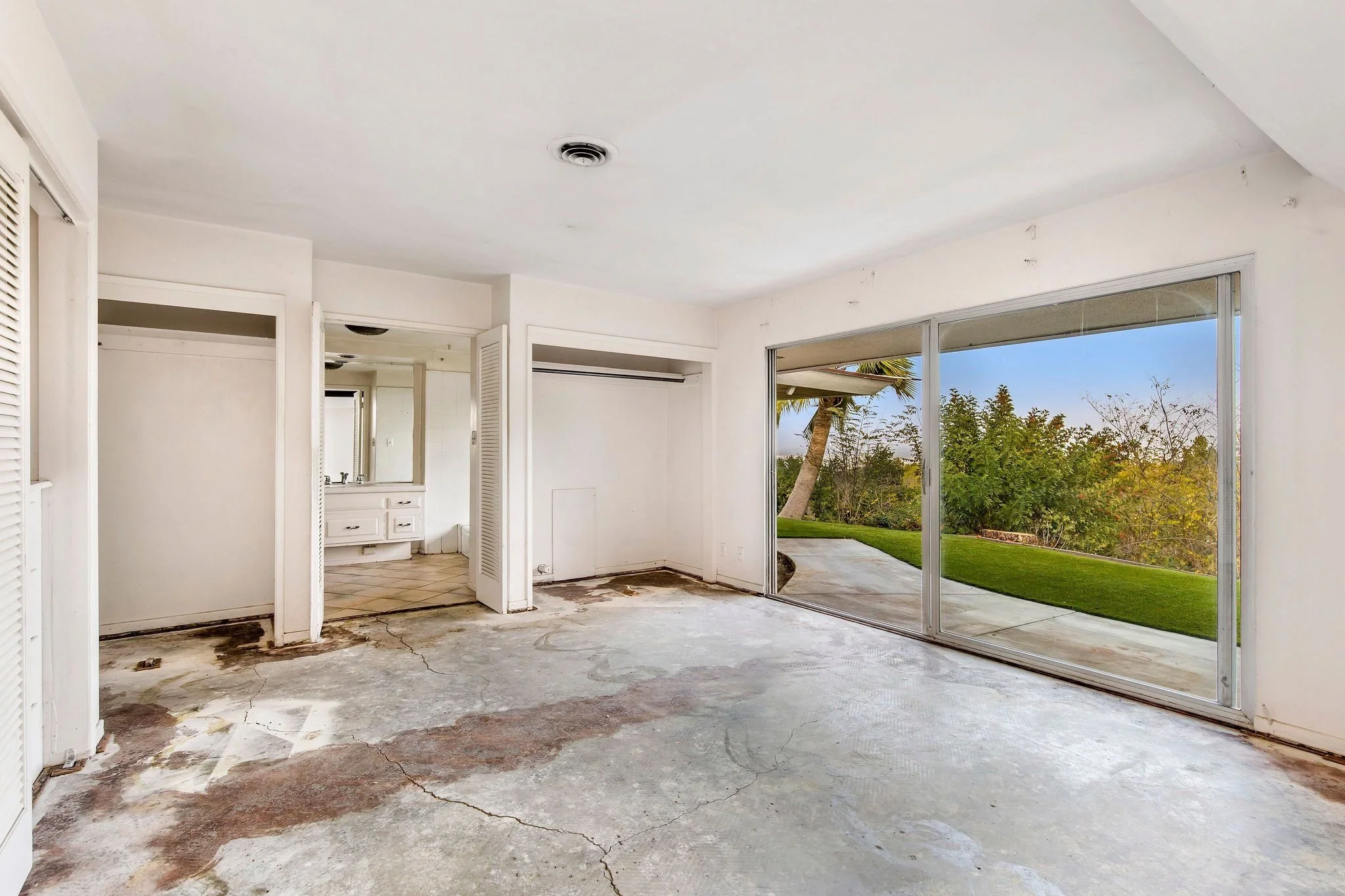Empty room with large sliding glass doors leading outside to a lawn with trees, visible flooring with some damage and cracks, and unfinished or damaged interior walls.