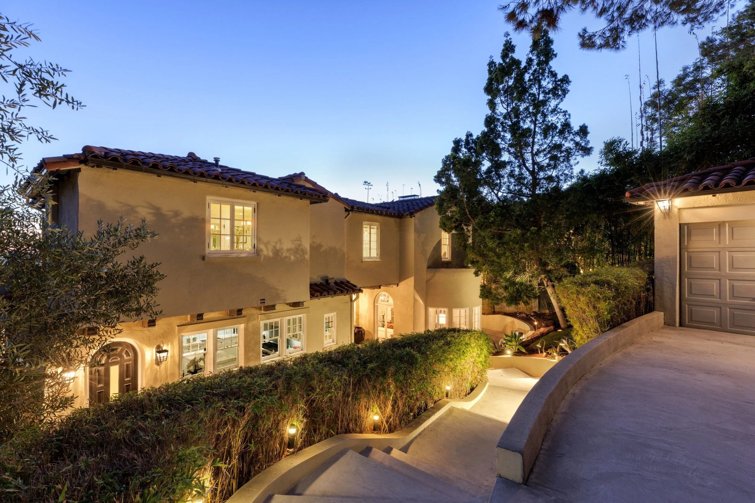 A residential house at dusk with a curved driveway, illuminated windows, garden lighting, and trees surrounding the house.
