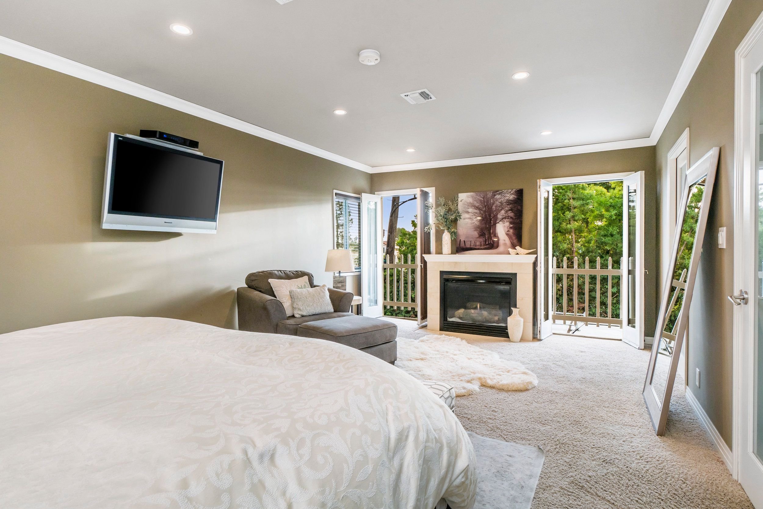 Living room with beige walls, a wall-mounted flat-screen TV, a gray armchair with a pillow, a fireplace, open doors leading to a balcony with trees outside, and a landscape painting above the fireplace.