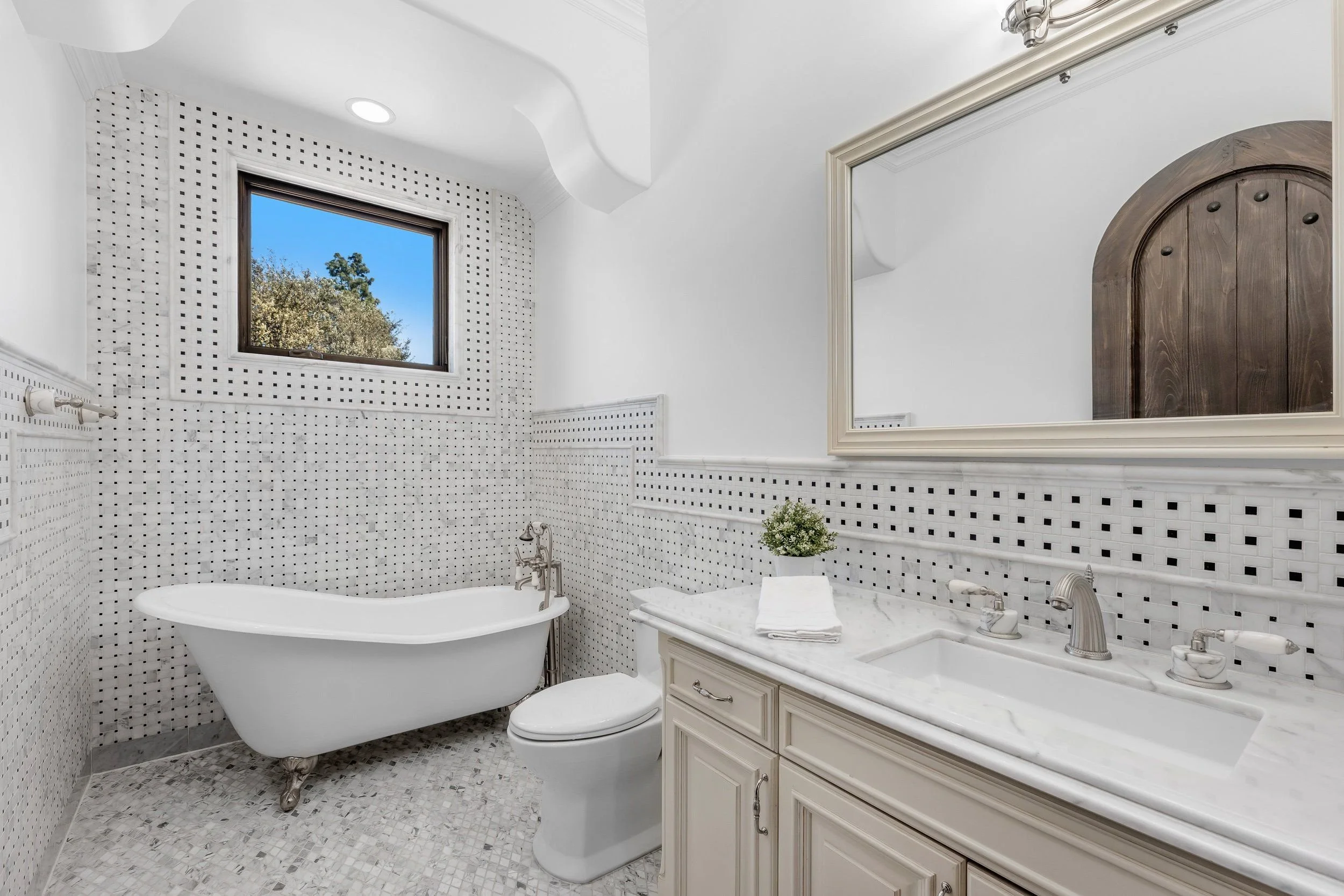 A white bathroom with a bathtub, toilet, and a cream-colored vanity with a marble countertop. The walls are tiled in small black and white squares, and there is a large mirror above the vanity. A window with a view of trees is above the bathtub, and 