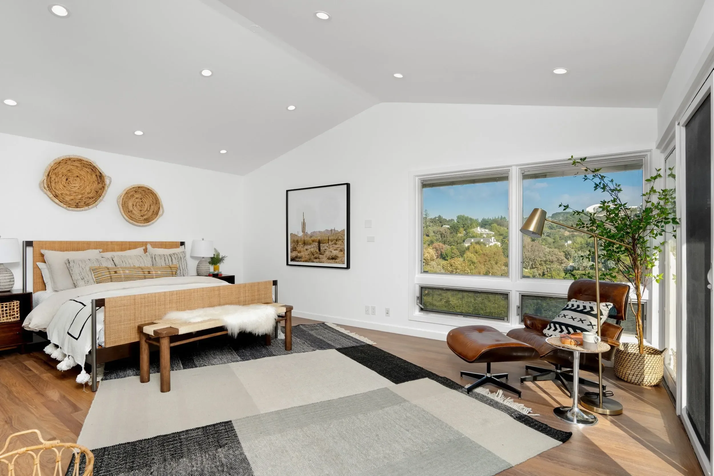 A bright bedroom with white walls and large windows overlooking a green hillside. The room features a wooden bed with white and beige bedding, a black and white area rug, a leather lounge chair with a side table, and decorative wall art including wov