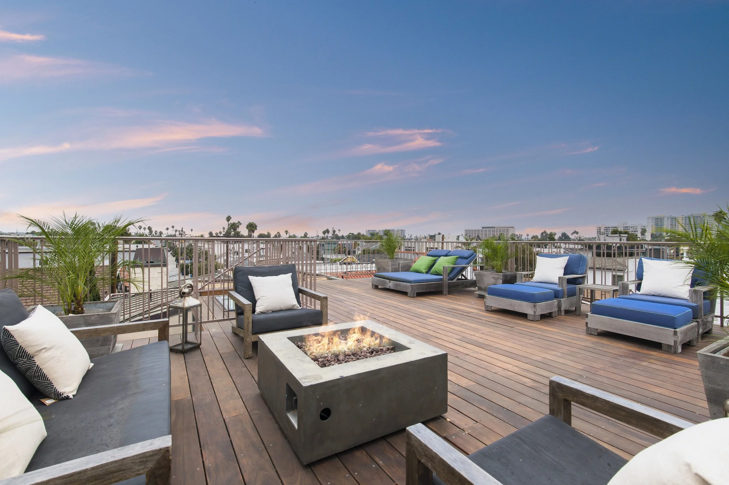 Rooftop patio with outdoor seating, a fire pit, potted plants, and views of city buildings and a sunset sky.