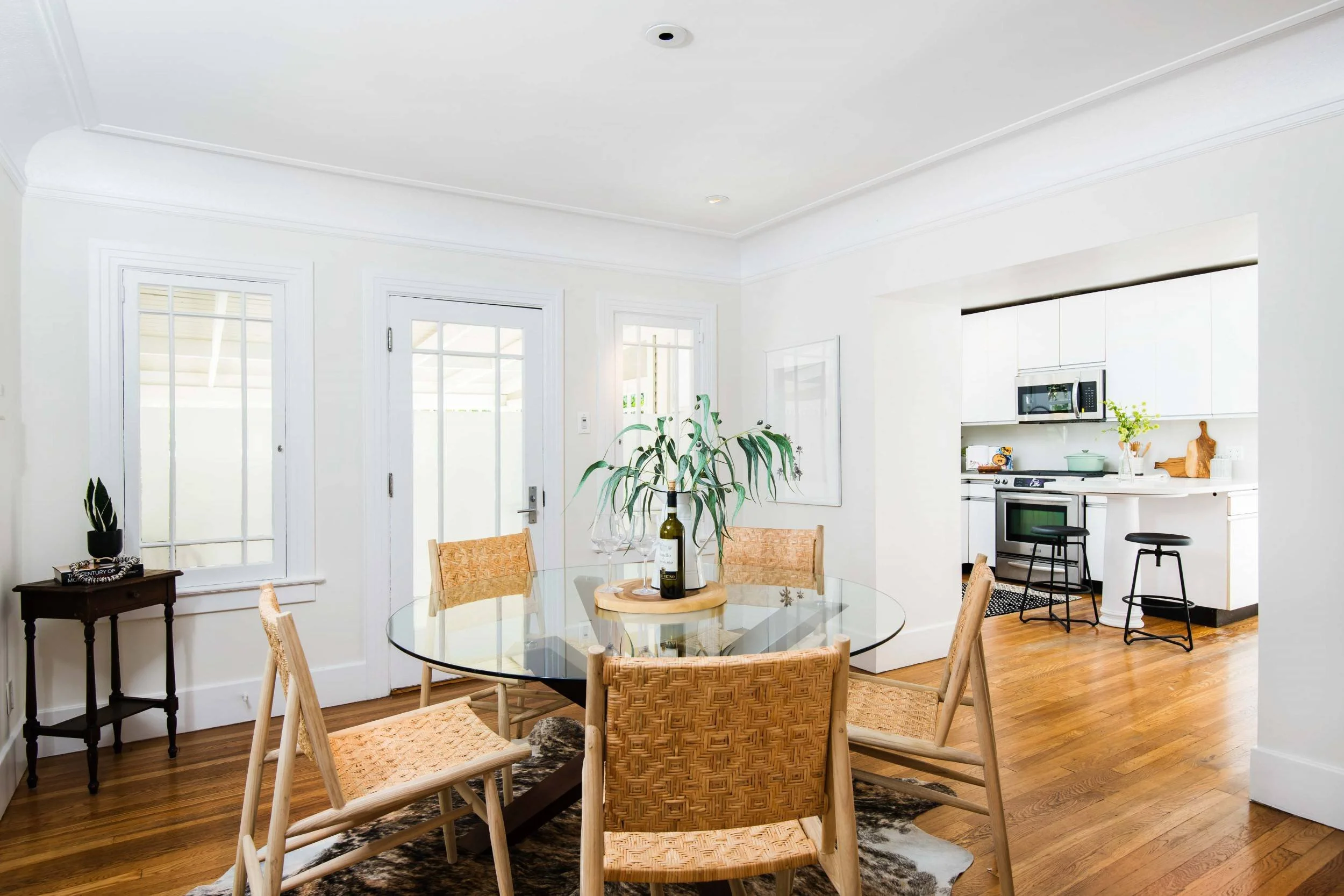 Bright dining area with round glass table, wooden chairs, and a side table with potted plants and books, adjacent to a kitchen with white cabinets and stainless steel appliances.