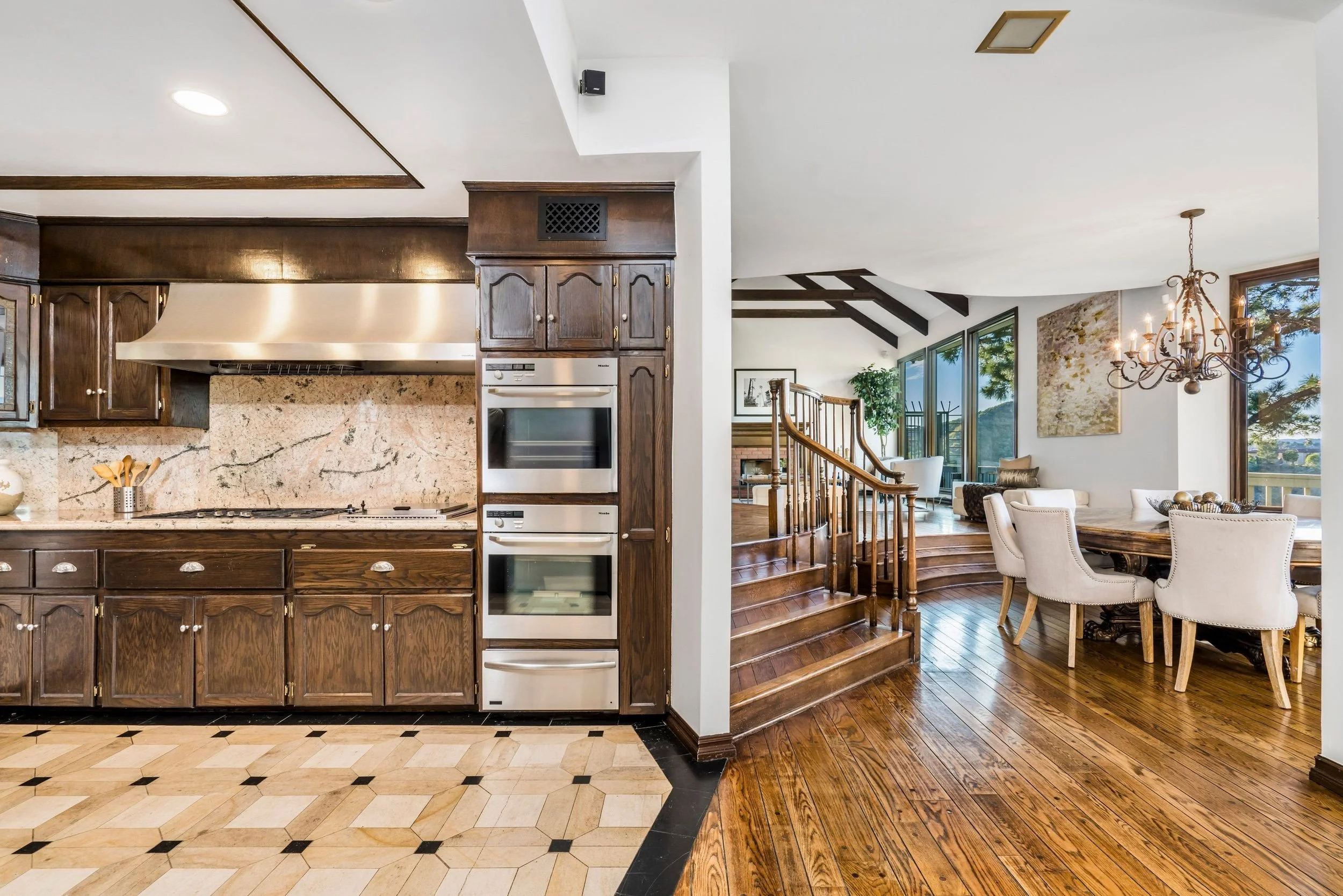 Interior view of a home with a kitchen and dining area, featuring wooden cabinets, a stone backsplash, a built-in oven, hardwood floors, a staircase, a chandelier, and large windows with a scenic view.