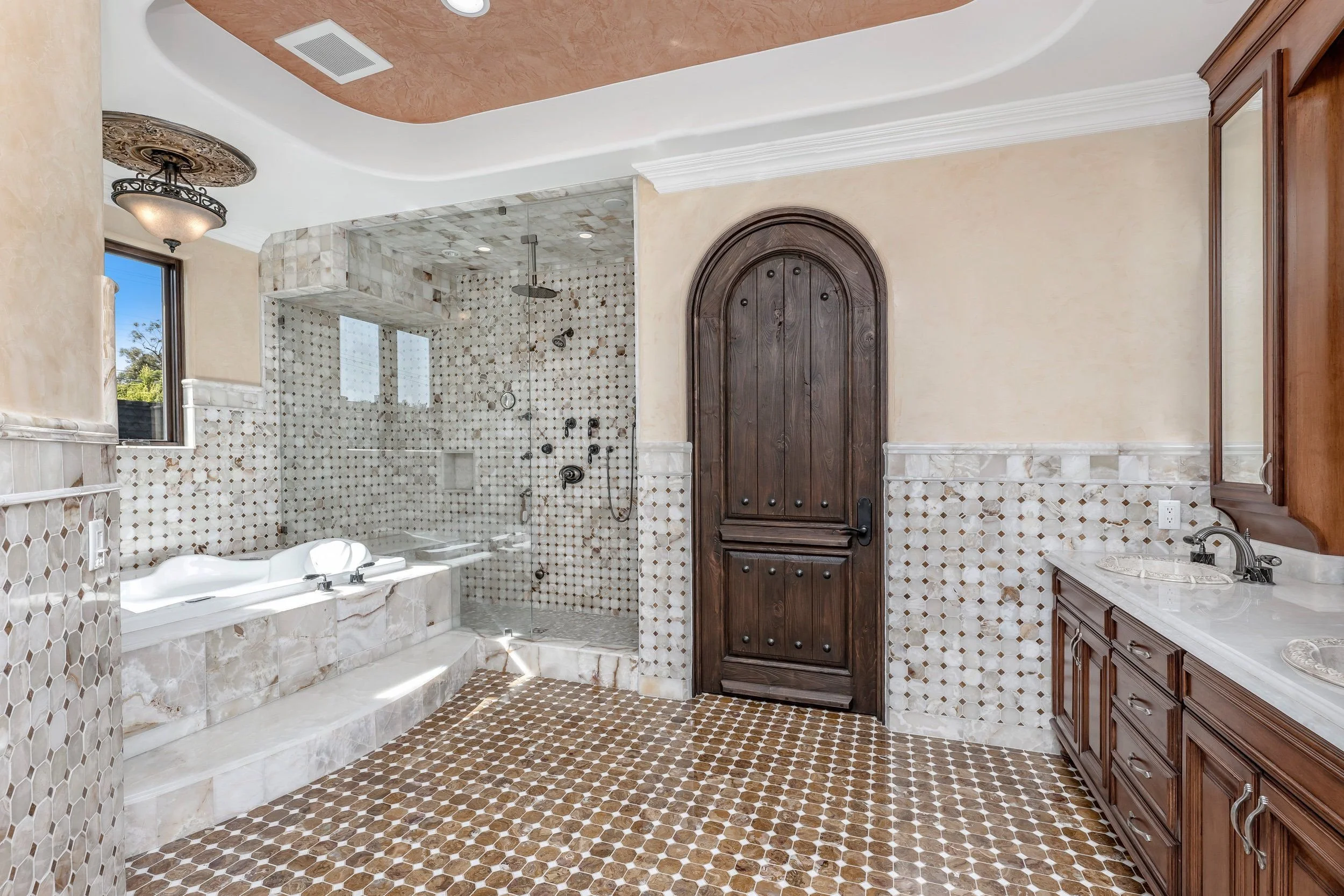 Luxury bathroom with walk-in shower, bathtub, wooden cabinets, marble countertops, and patterned tile flooring.