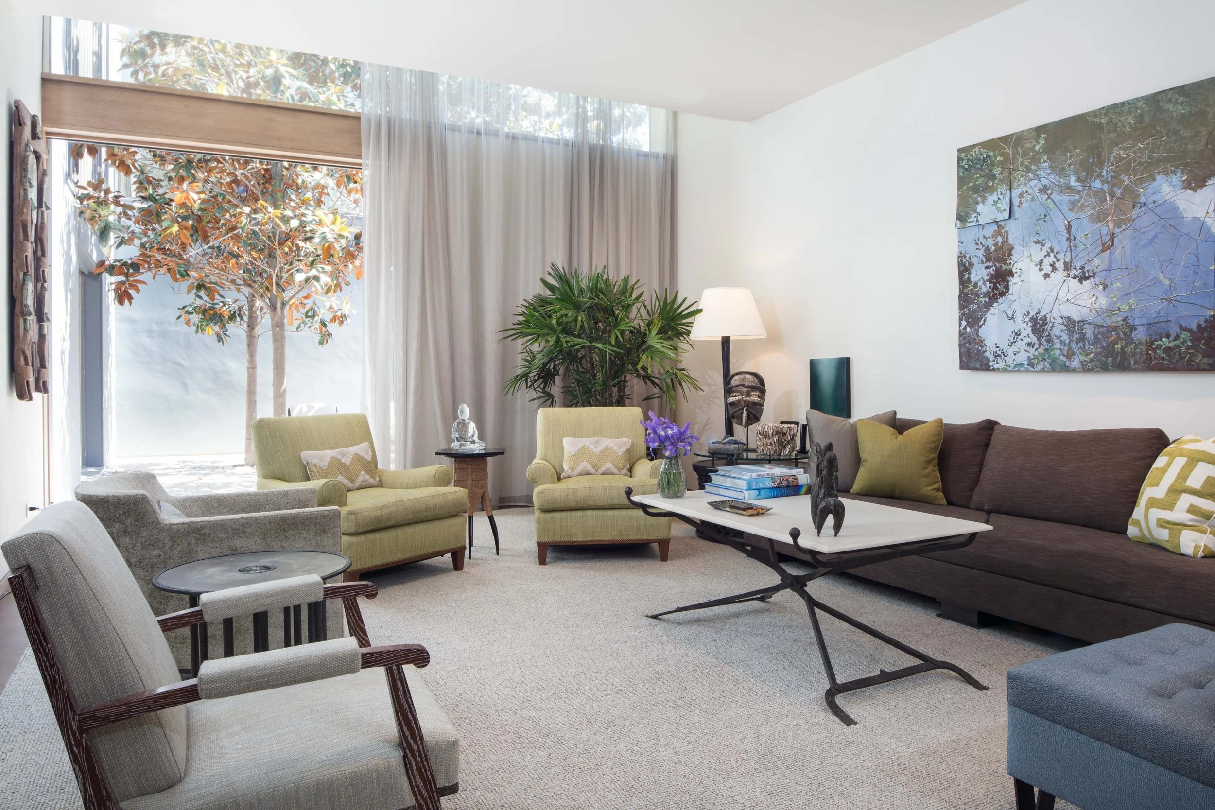 Bright living room with large window, sheer curtains, and a view of a tree outside. Contains green and brown upholstered chairs, a brown sofa with pillows, a coffee table with books, decorative sculptures, and houseplants, with artwork hanging on the wall.