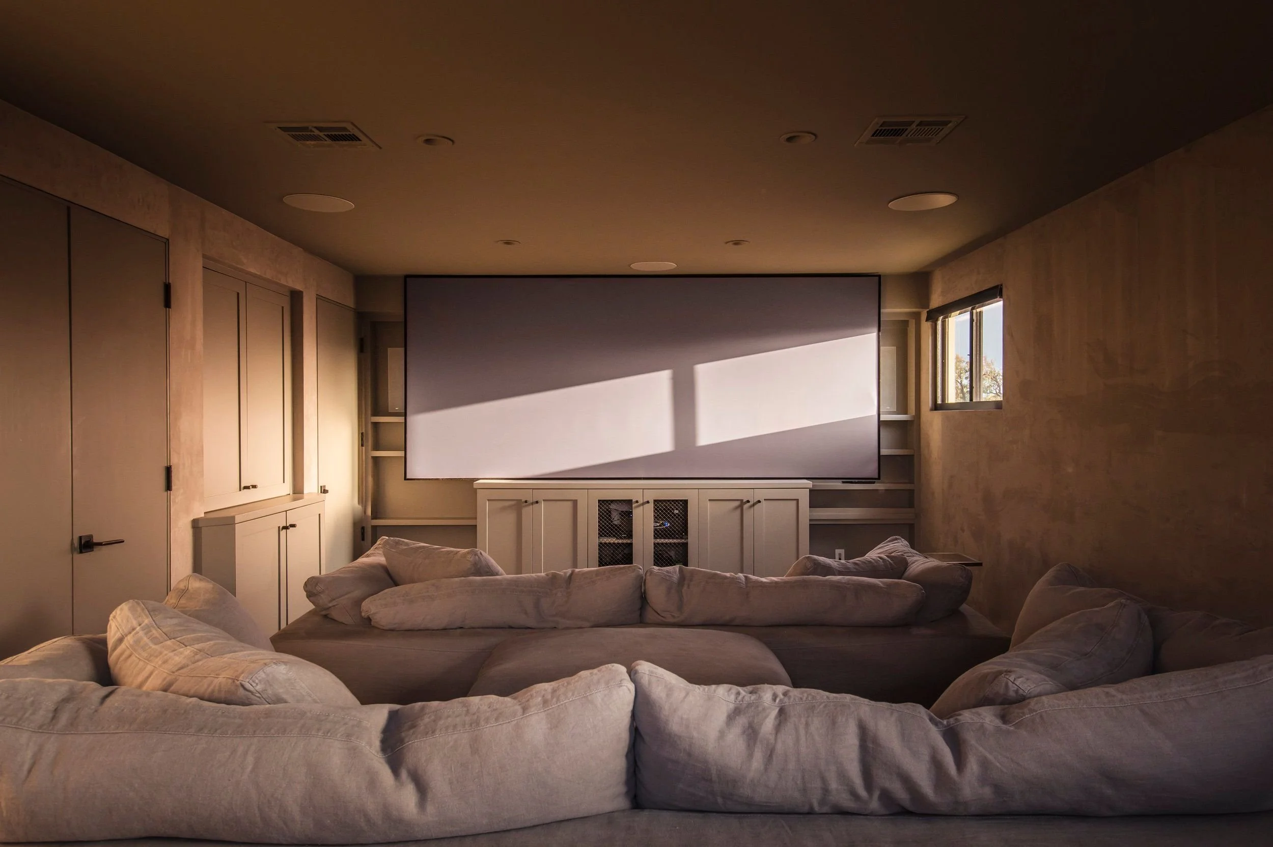 Living room with beige sectional sofa and large flat-screen TV mounted on the wall, sunlight coming through window, built-in cabinets, and textured beige walls.