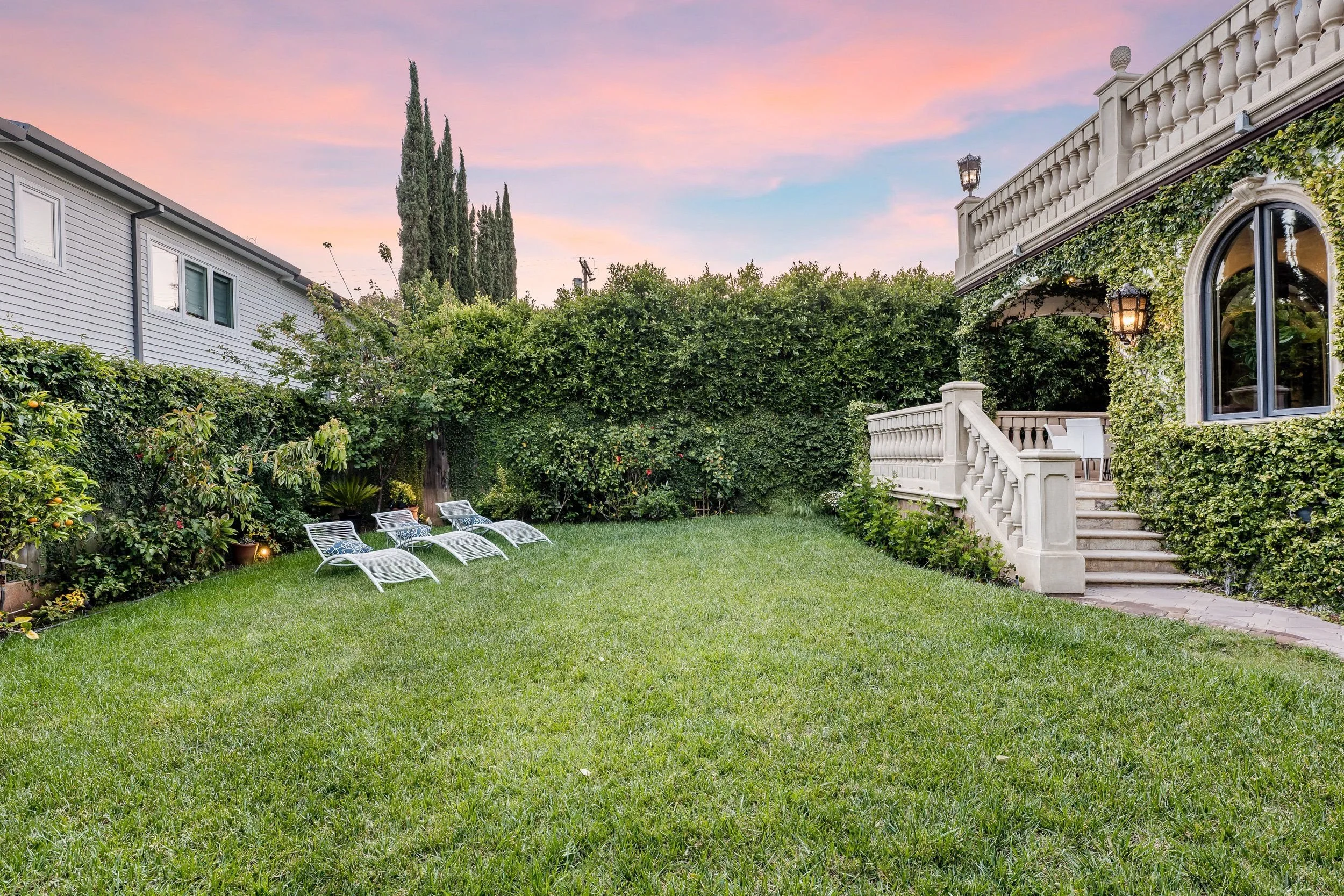 Backyard with green grass, several lounge chairs, surrounded by lush bushes and trees, and a white house with a patio on the right, during sunset with pastel pink and blue sky.