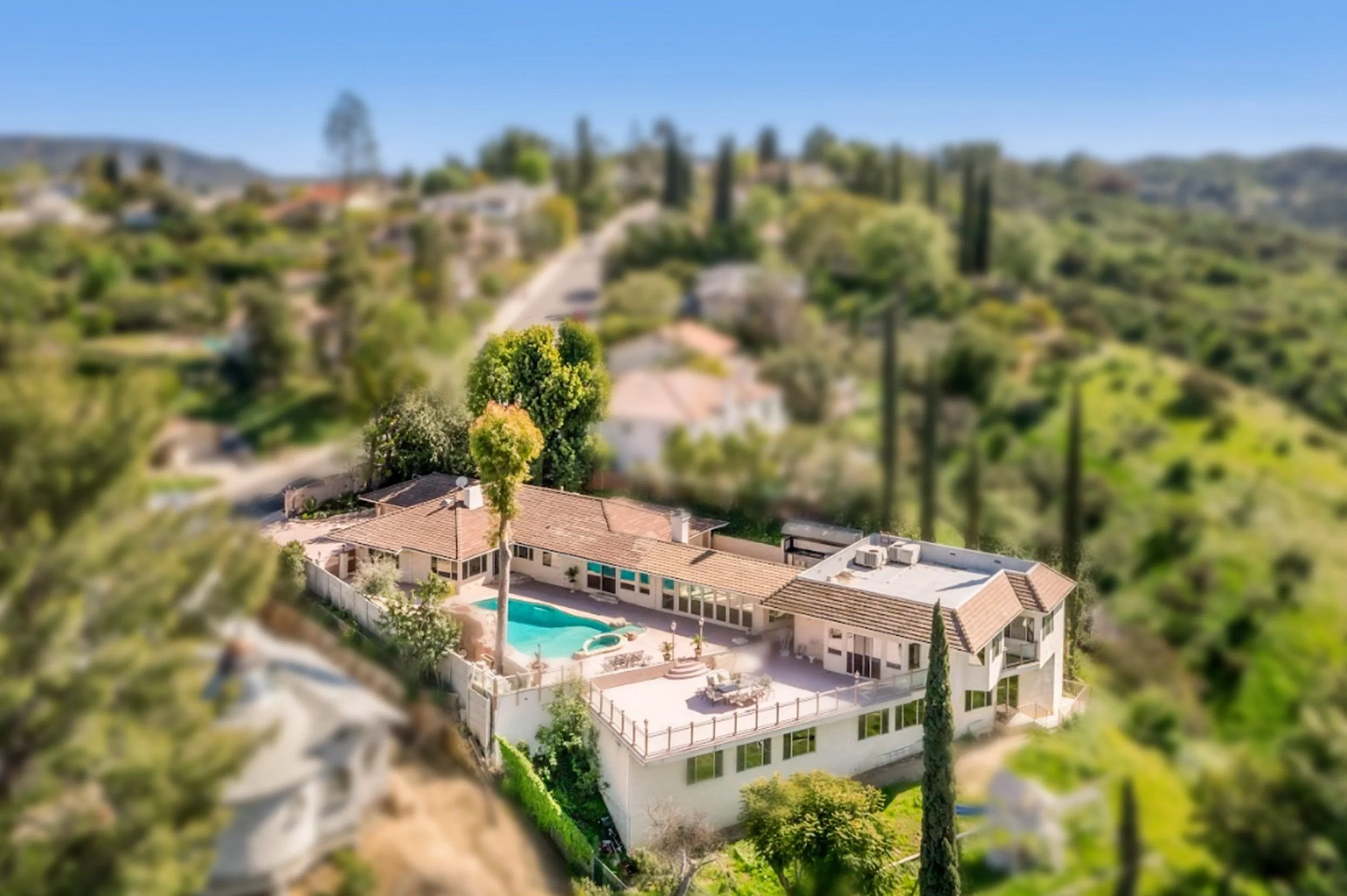 Aerial view of a hillside residential area with houses, trees, and a swimming pool, under a clear blue sky.
