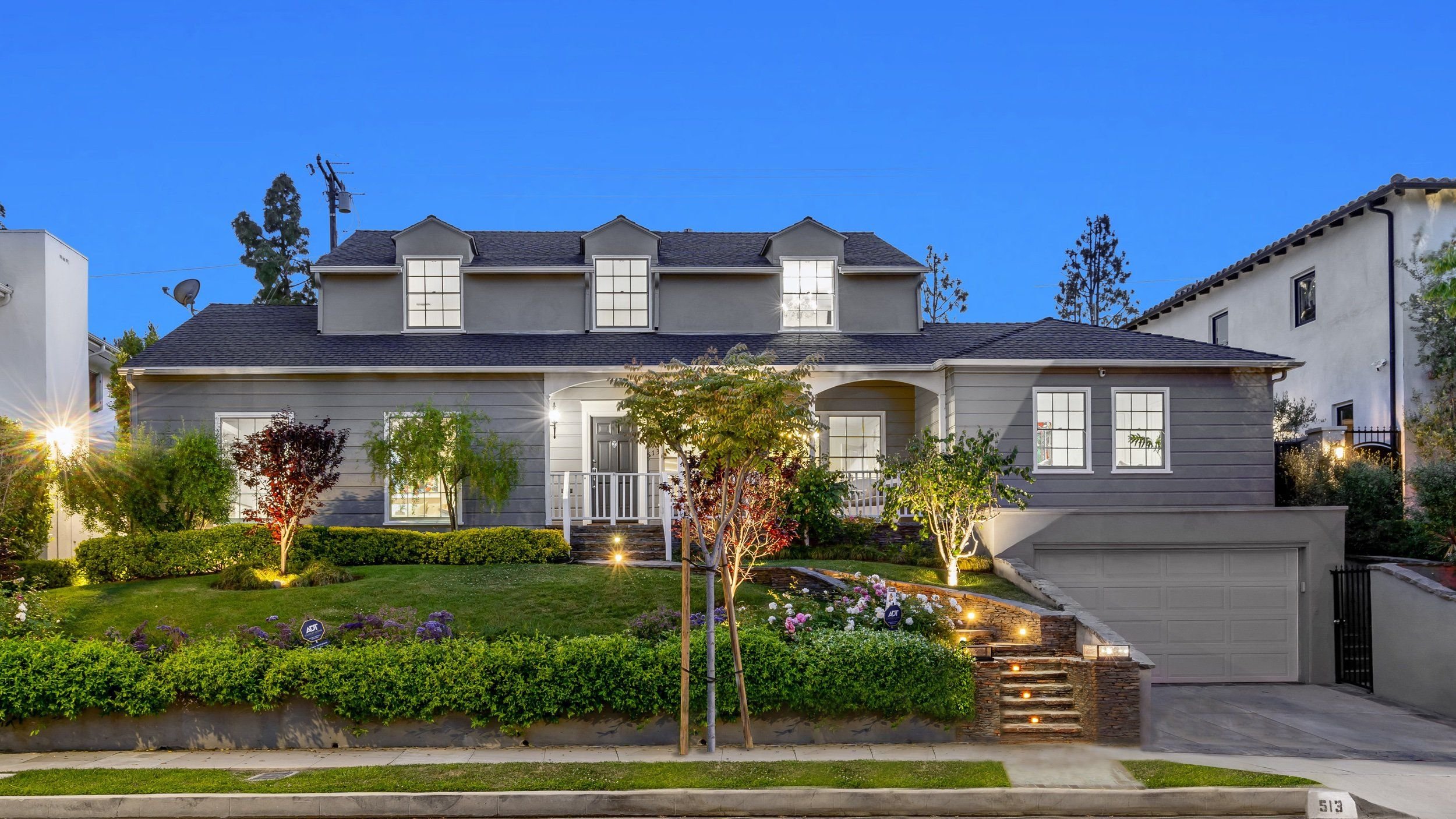 A modern two-story house with gray siding, multiple windows, and a front porch with a small railing. The landscaped front yard has trees, shrubs, and flowers, illuminated by outdoor lighting. The driveway leads to a garage under the house, surrounded