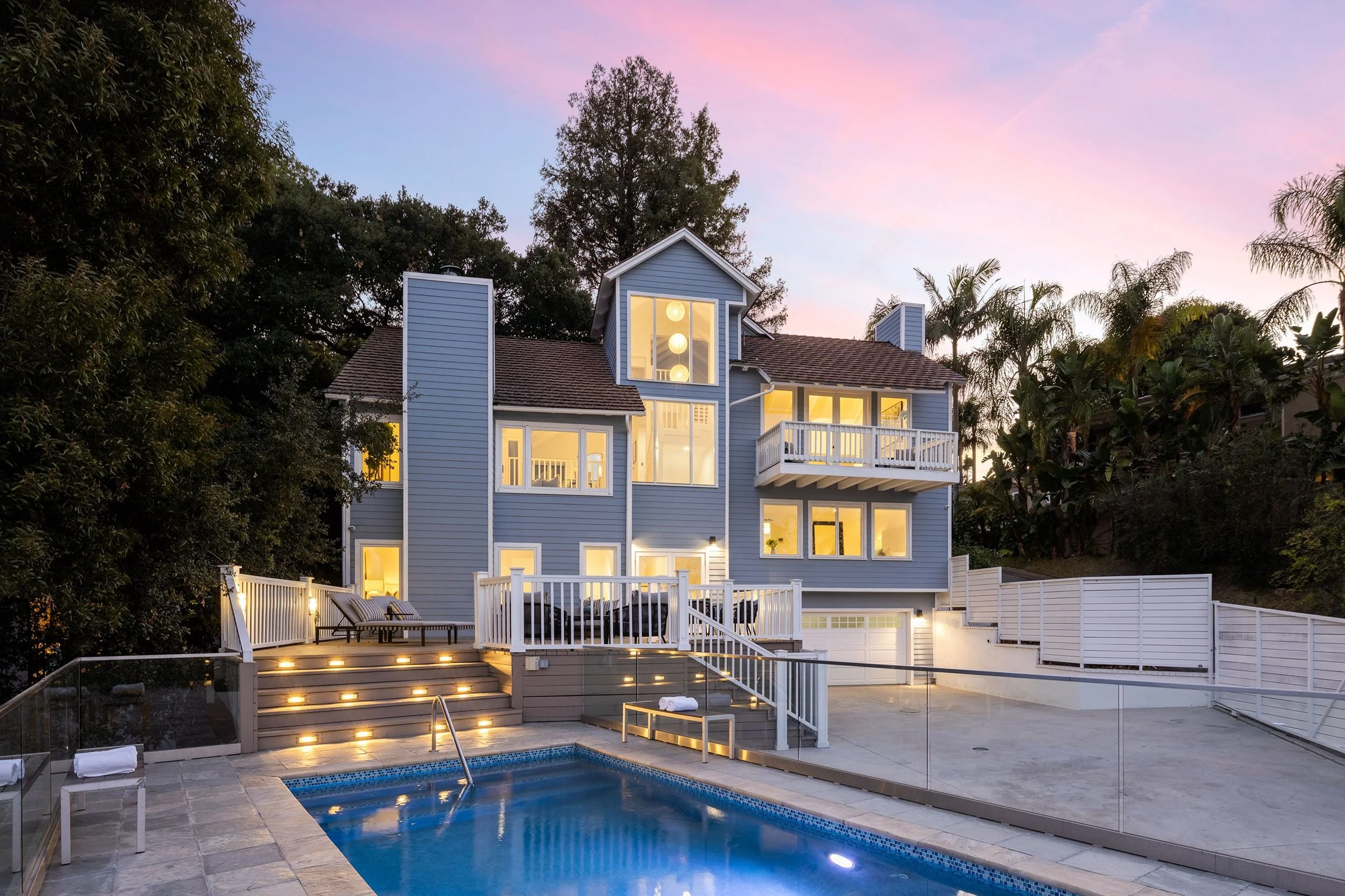 A two-story blue house with large windows, a balcony, and a backyard with a swimming pool. The house is illuminated from within, and the sky shows a pink and purple sunset. There are plants and trees surrounding the house, with a patio and outdoor fu