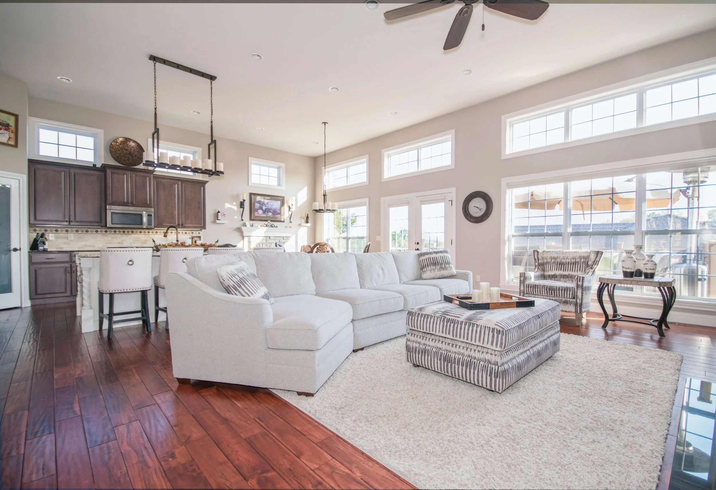 Living room with white sectional sofa, striped ottoman, large windows, hardwood floors, fireplace, wall clock, and kitchen area in background.