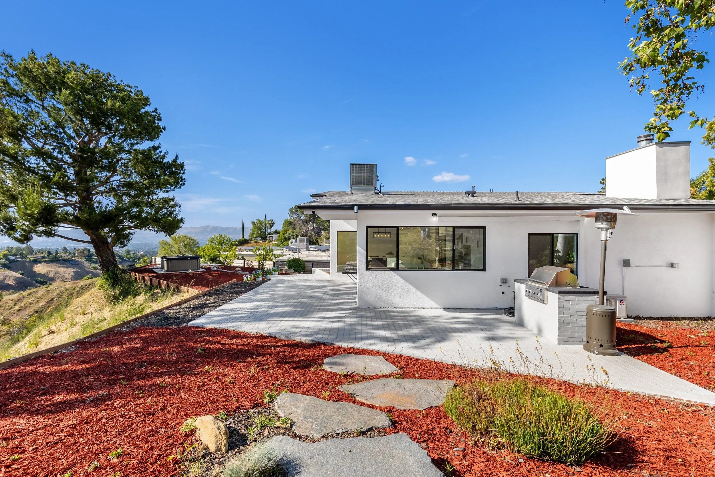 View of a modern white house with large windows in a sunny outdoor setting. The house has a small patio area with outdoor furniture and a barbecue grill. The foreground features a landscaped yard with red mulch, small rocks, green plants, and a large