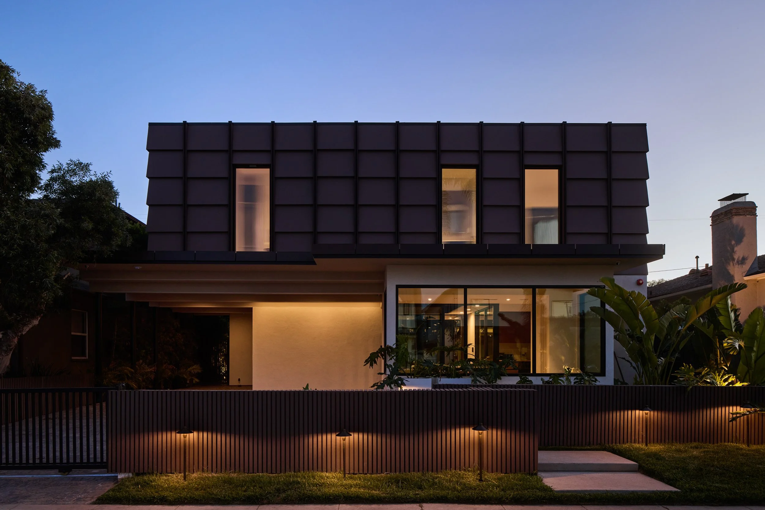 Modern two-story house with large windows, dark exterior panels, a gated front yard, trees, and outdoor lighting at dusk.