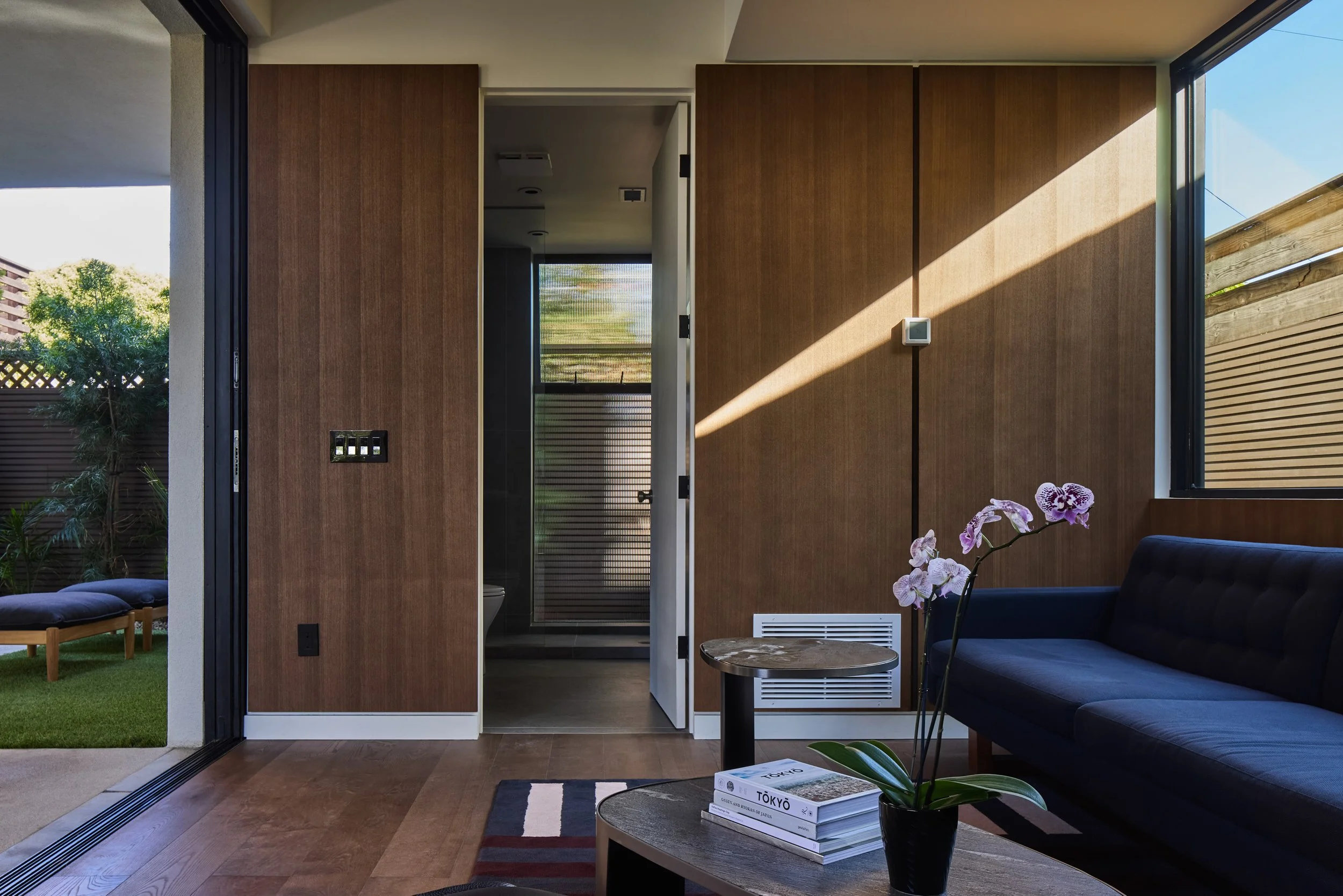 Modern living room with a navy sofa, a wooden coffee table with books and a potted orchid, wood-paneled wall, open glass door revealing a garden, and sunlight casting shadow patterns.