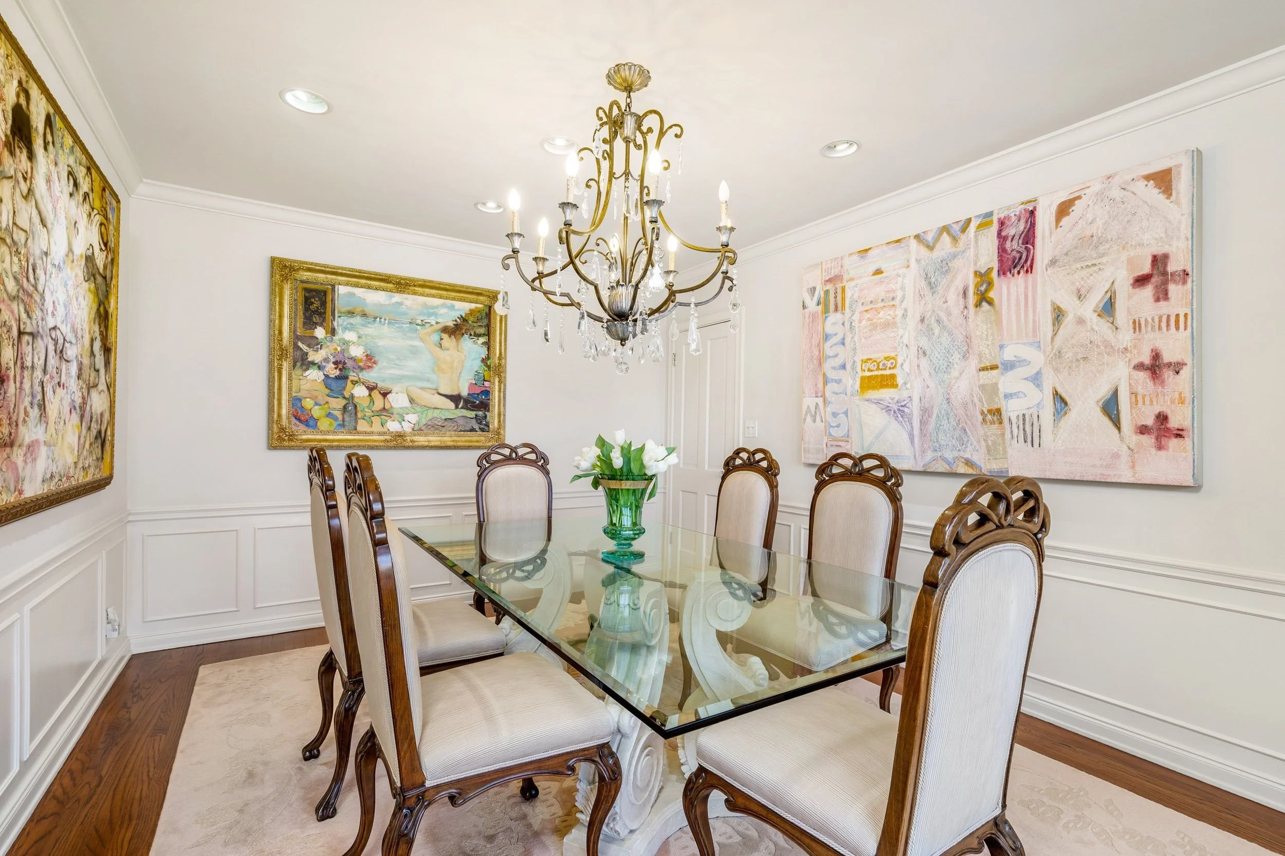 A dining room with a glass-top table, cream upholstered chairs, a chandelier, and colorful artwork on the walls.