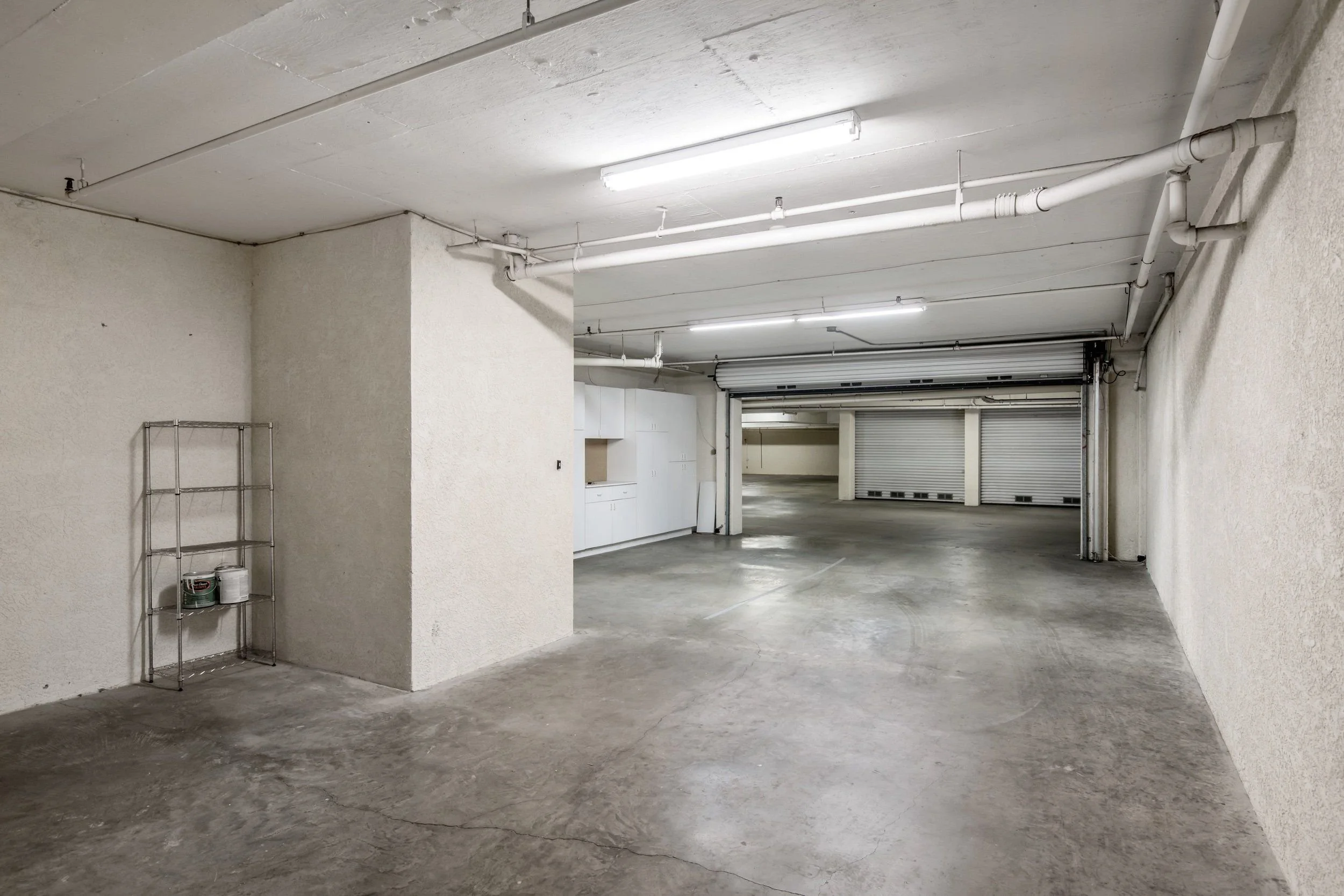 Empty underground parking garage with concrete floors and white walls, fluorescent lighting, and a partially open roll-up garage door.