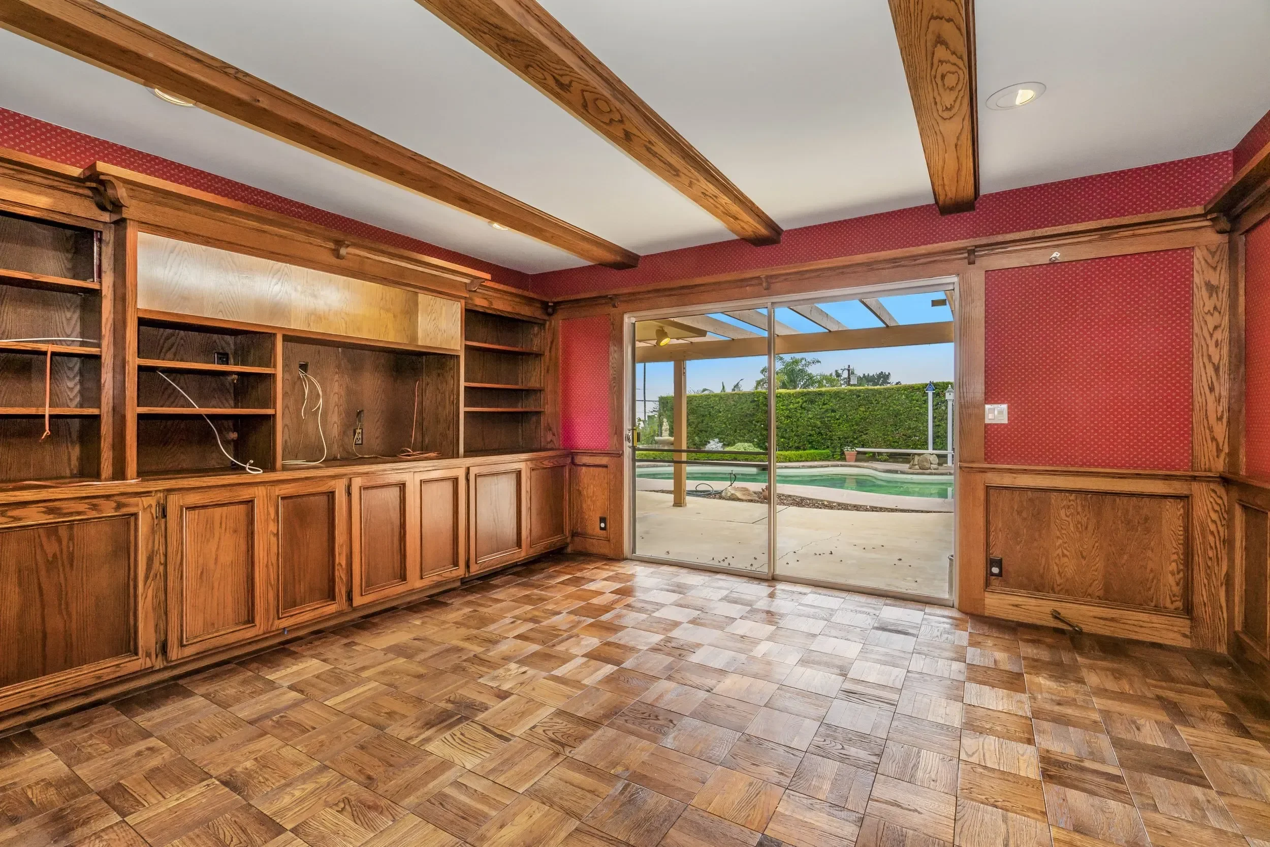 Interior view of a room with wooden cabinets and shelves, a sliding glass door leading outside to a patio with a swimming pool, and a view of hedges and trees in the distance.