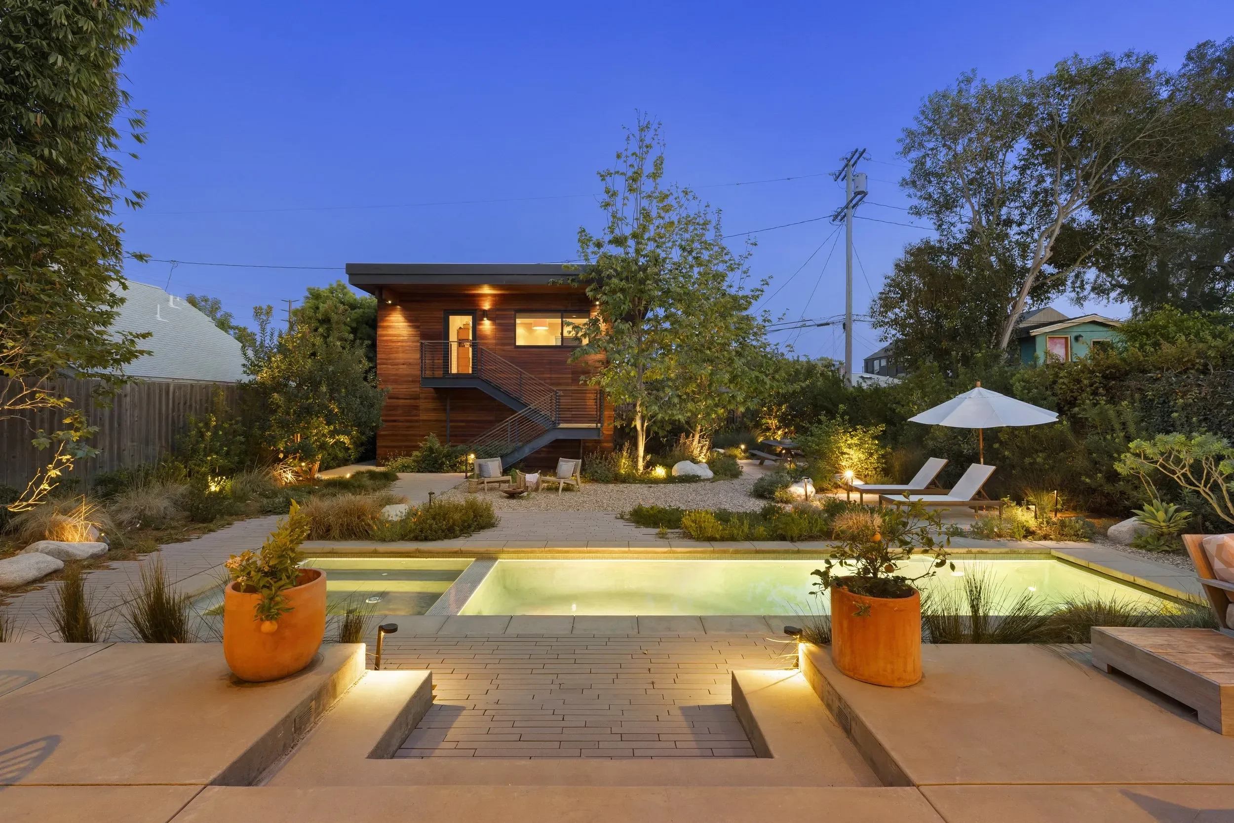 Modern backyard with a swimming pool surrounded by potted plants, outdoor seating, trees, and a wooden house under a clear evening sky.