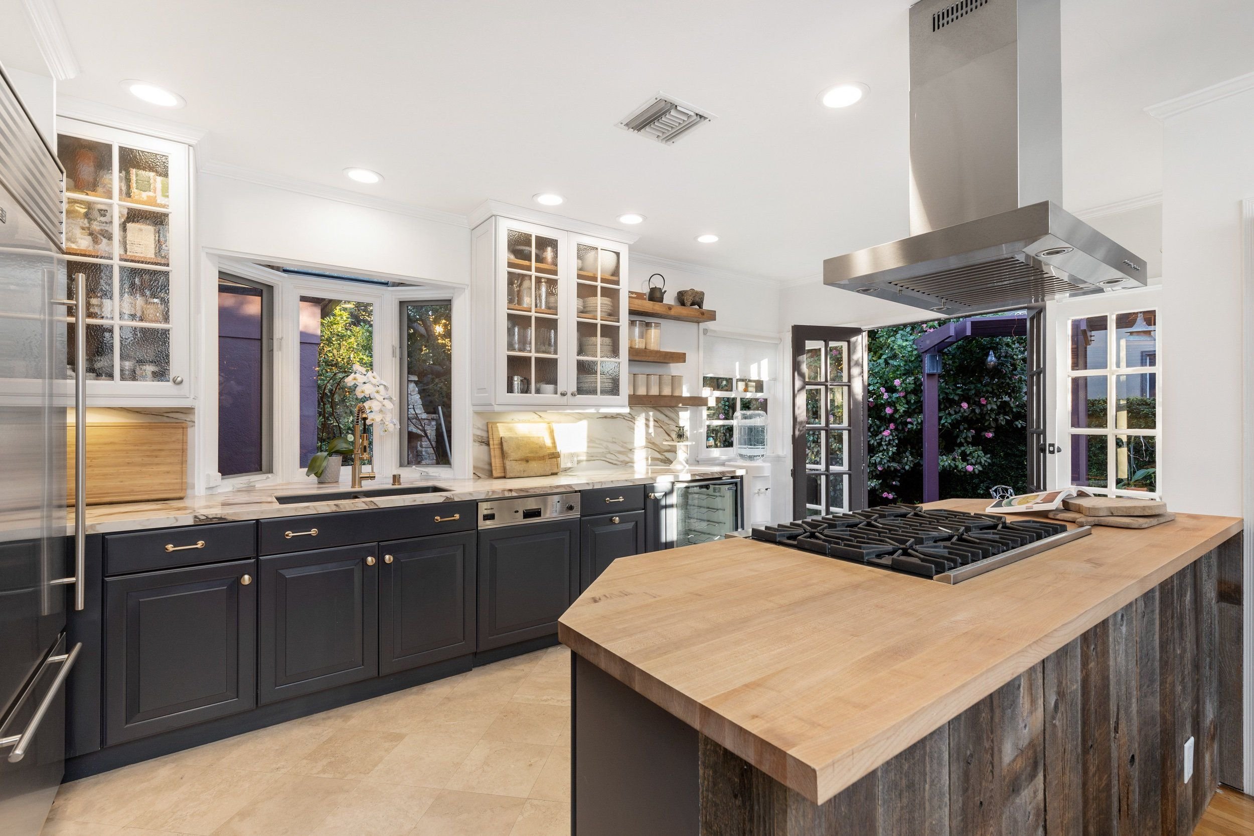 Contemporary kitchen with white cabinets, black lower cabinets, a large wooden kitchen island, stainless steel range hood, stovetop, and open shelves with decor items and dishes. There are multiple windows letting in natural light and a door leading 