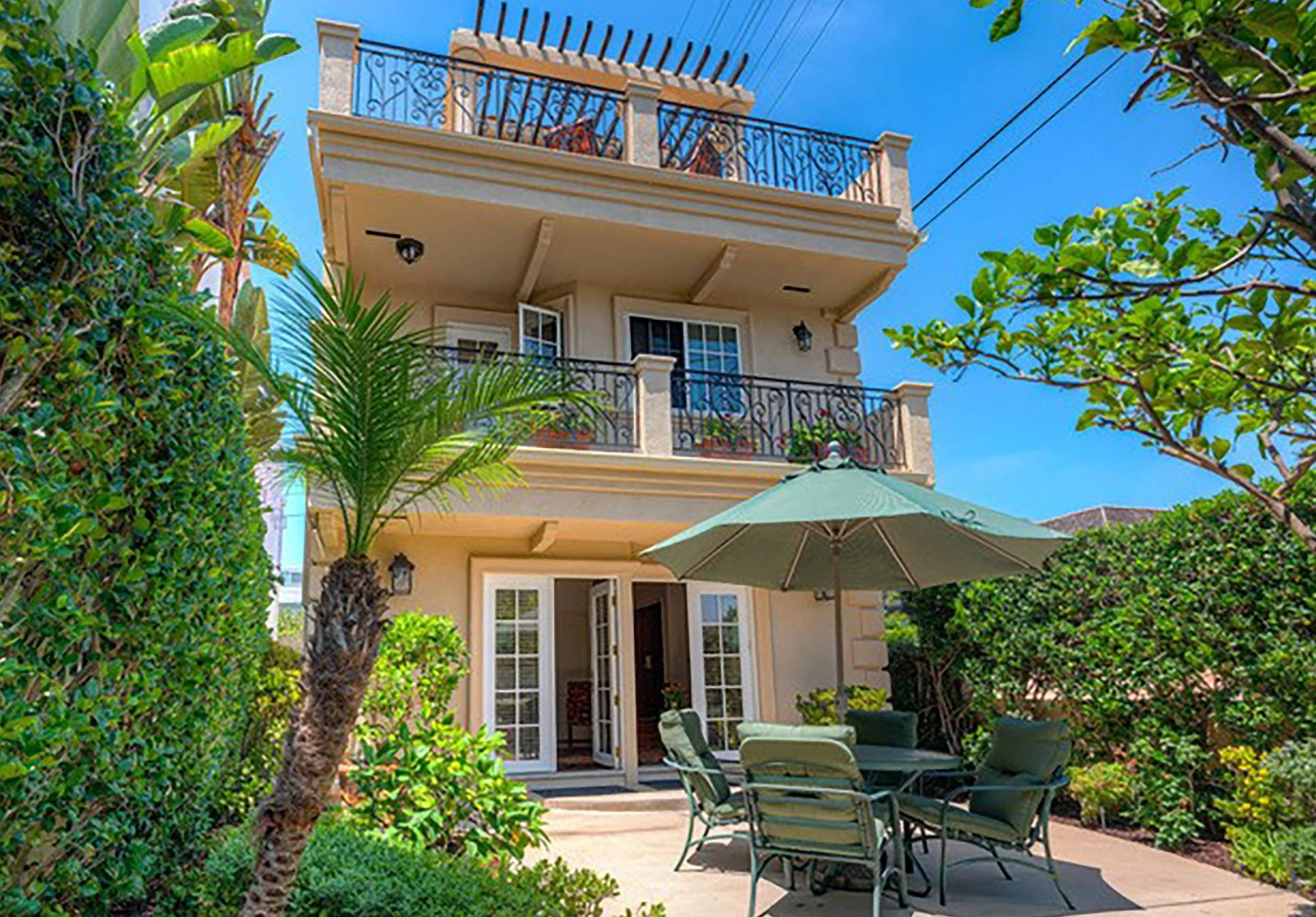 A residential house with a beige exterior, multiple balconies with black wrought iron railings, a patio with outdoor furniture, a large green patio umbrella, and lush trees and bushes surrounding the area under a clear blue sky.