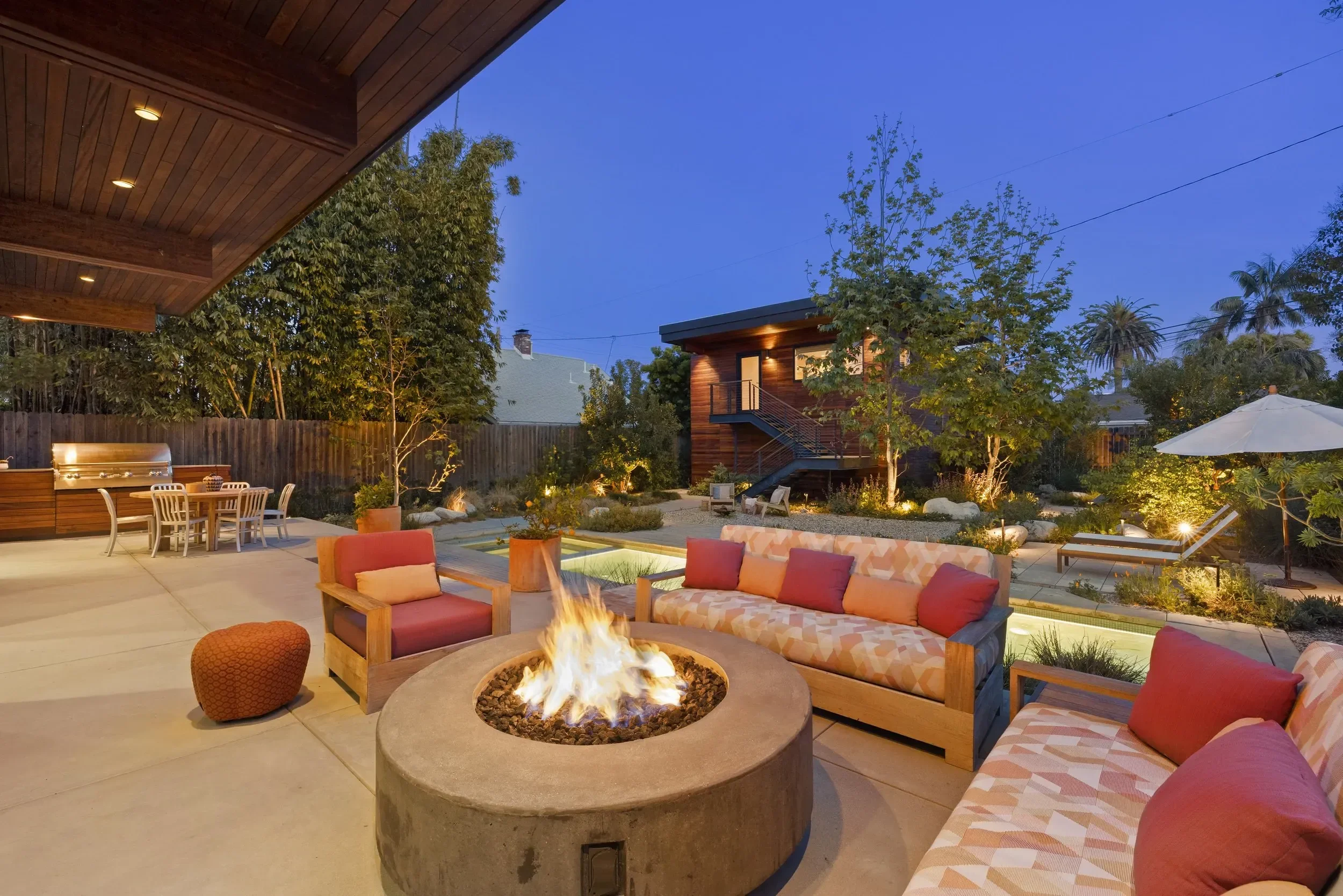 A backyard patio area at dusk with outdoor furniture, a fire pit with flames, trees, bushes, a wooden house with stairs, and an umbrella in the background.