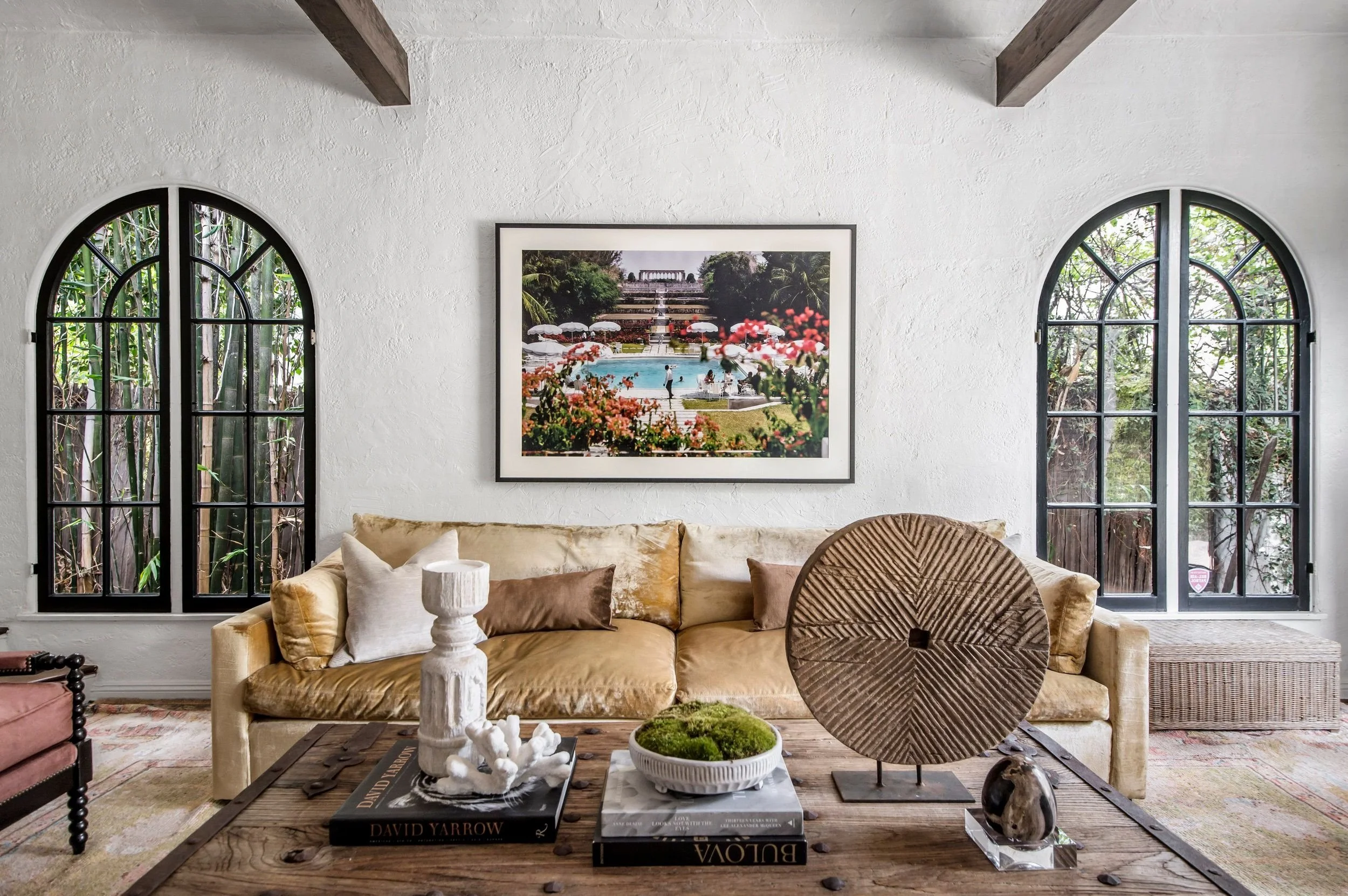 Living room with large beige sofa, wooden coffee table, and two arched windows with greenery outside. Art photograph of a pool and garden wall hangs on white wall.