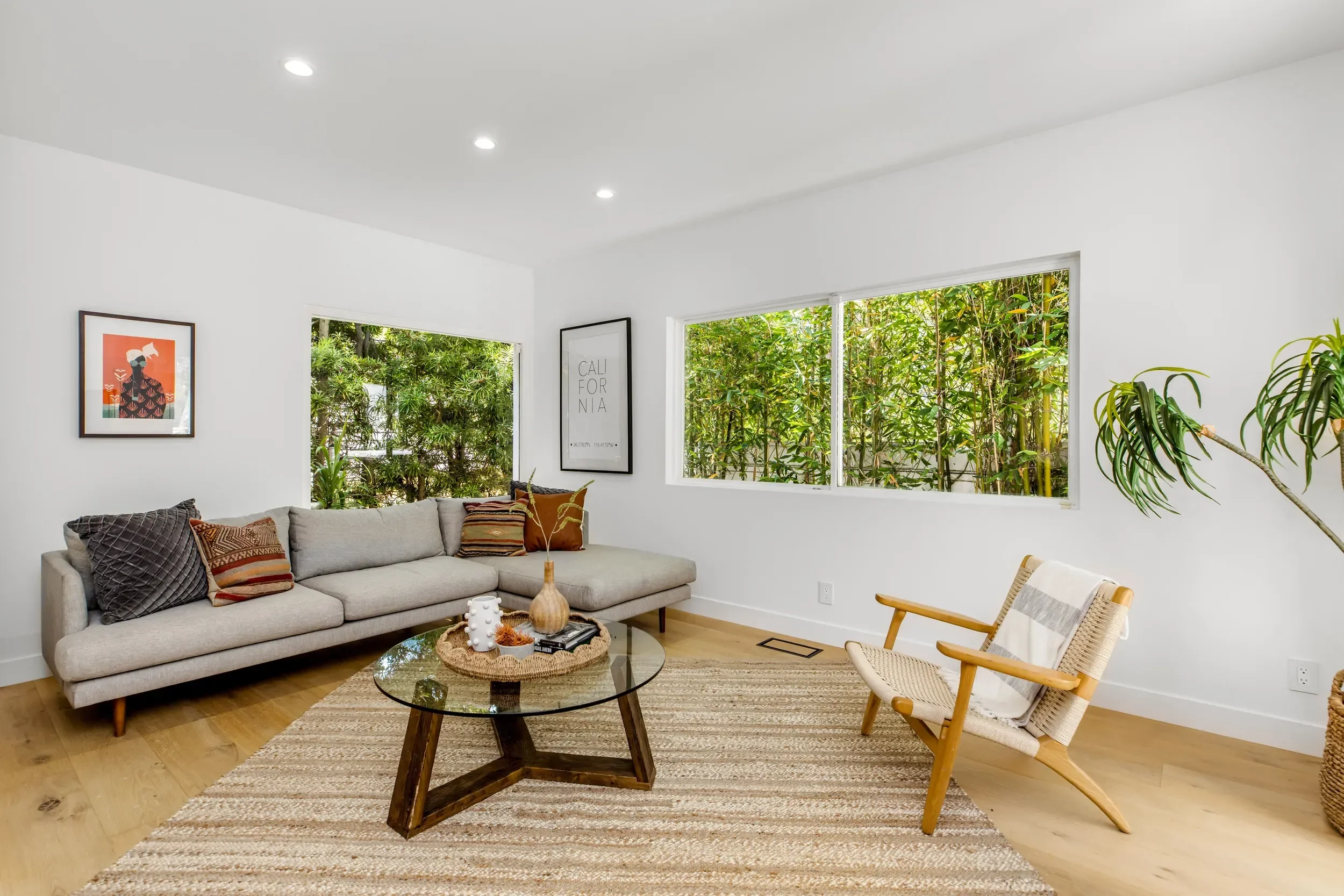 Living room with a beige sofa, colorful pillows, a glass coffee table with books and decor, a wooden armchair, a large beige area rug, white walls with framed artwork, and large windows with greenery outside.