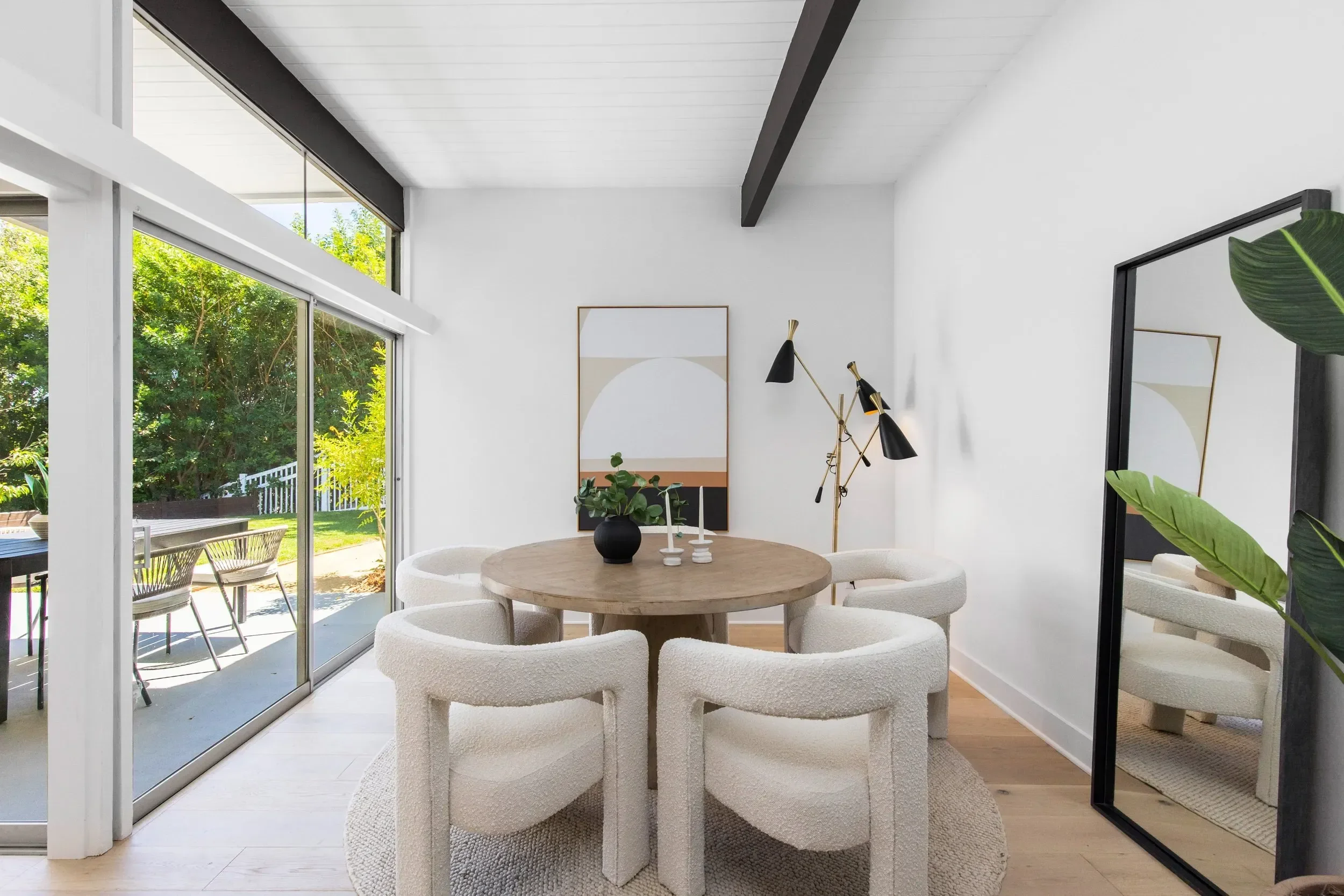 Modern dining room with white walls, large sliding glass doors to an outdoor patio, round wooden table with six textured white chairs, black and gold floor lamp, wall art, and green plants.