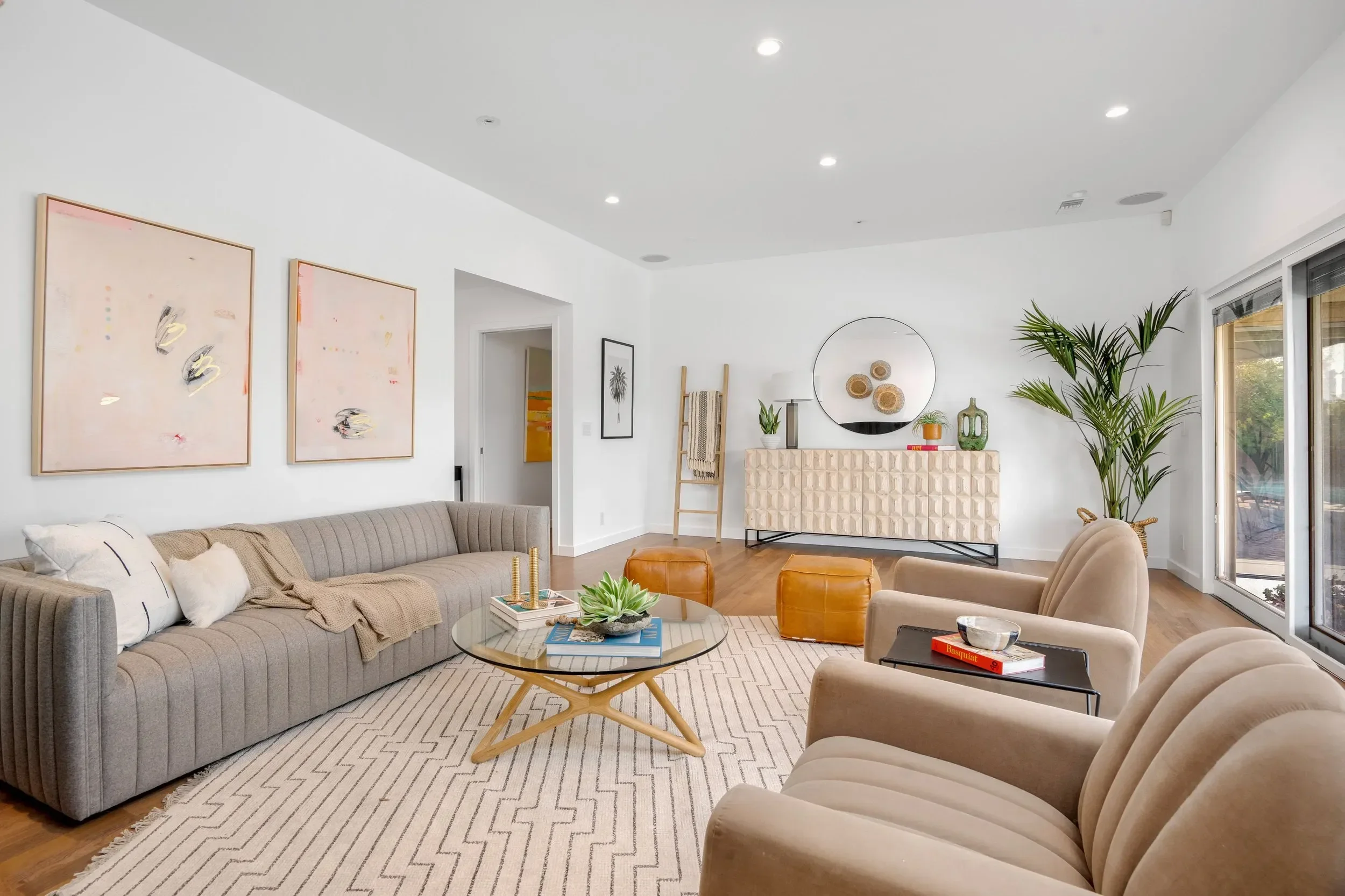 Modern living room with beige and gray furniture, abstract wall art, a large leafy plant, and sliding glass doors.