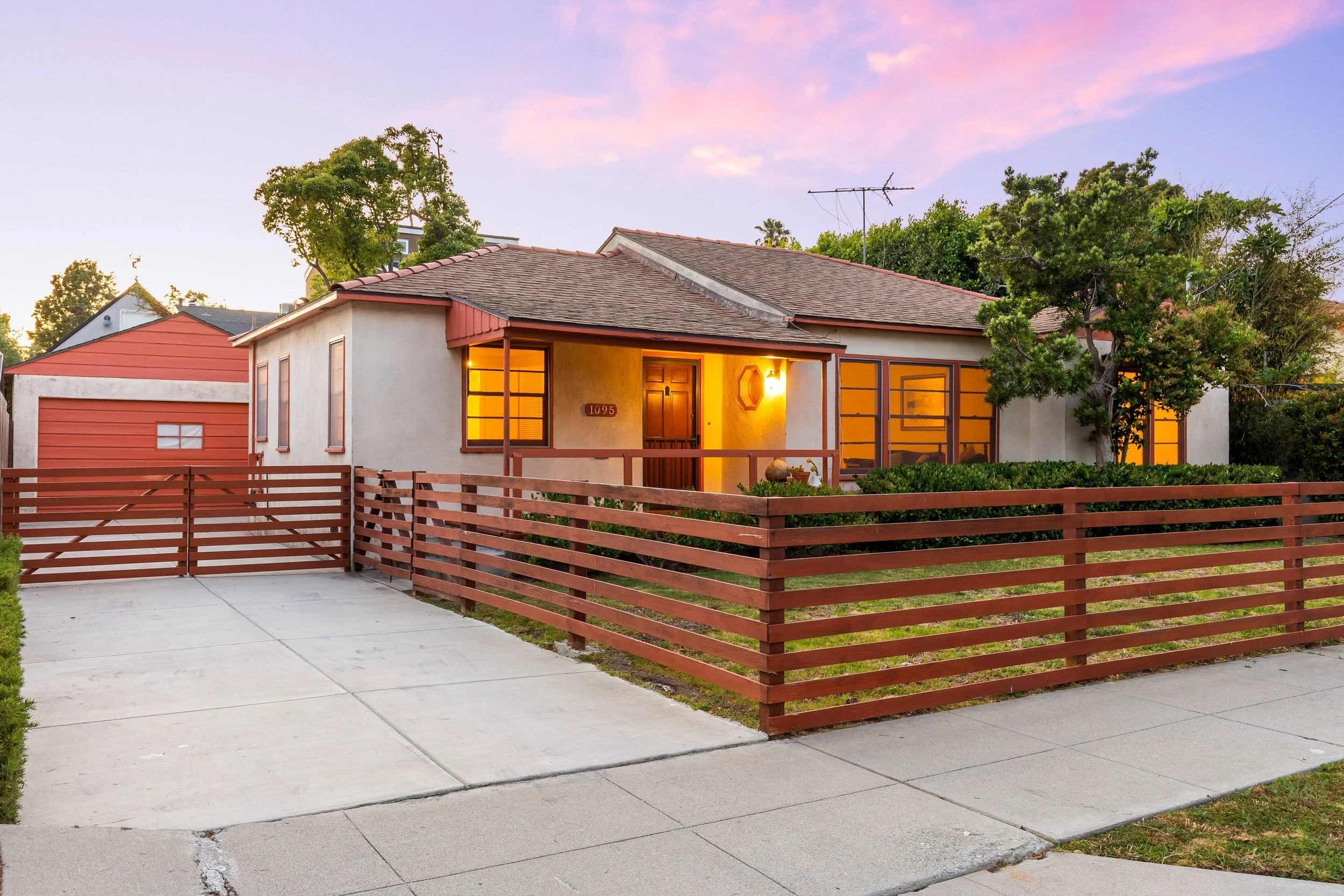 A single-story house with a red-tiled roof and beige exterior walls, illuminated from inside, with a wooden fence and sidewalk in front, surrounded by trees and a clear sky at sunset.