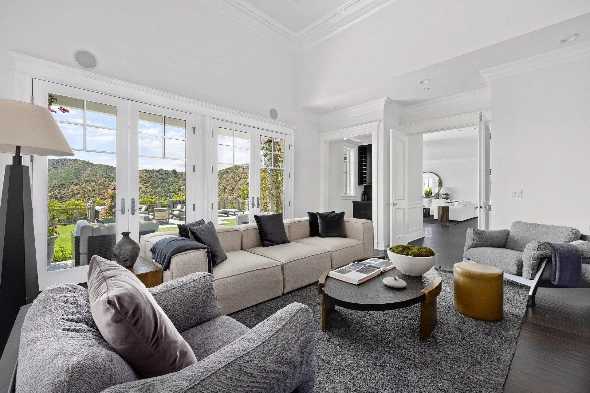 Living room with large glass doors and scenic mountain view, white walls and ceiling, gray and beige furniture, black and white pillows, modern decor, and a dark wood coffee table.