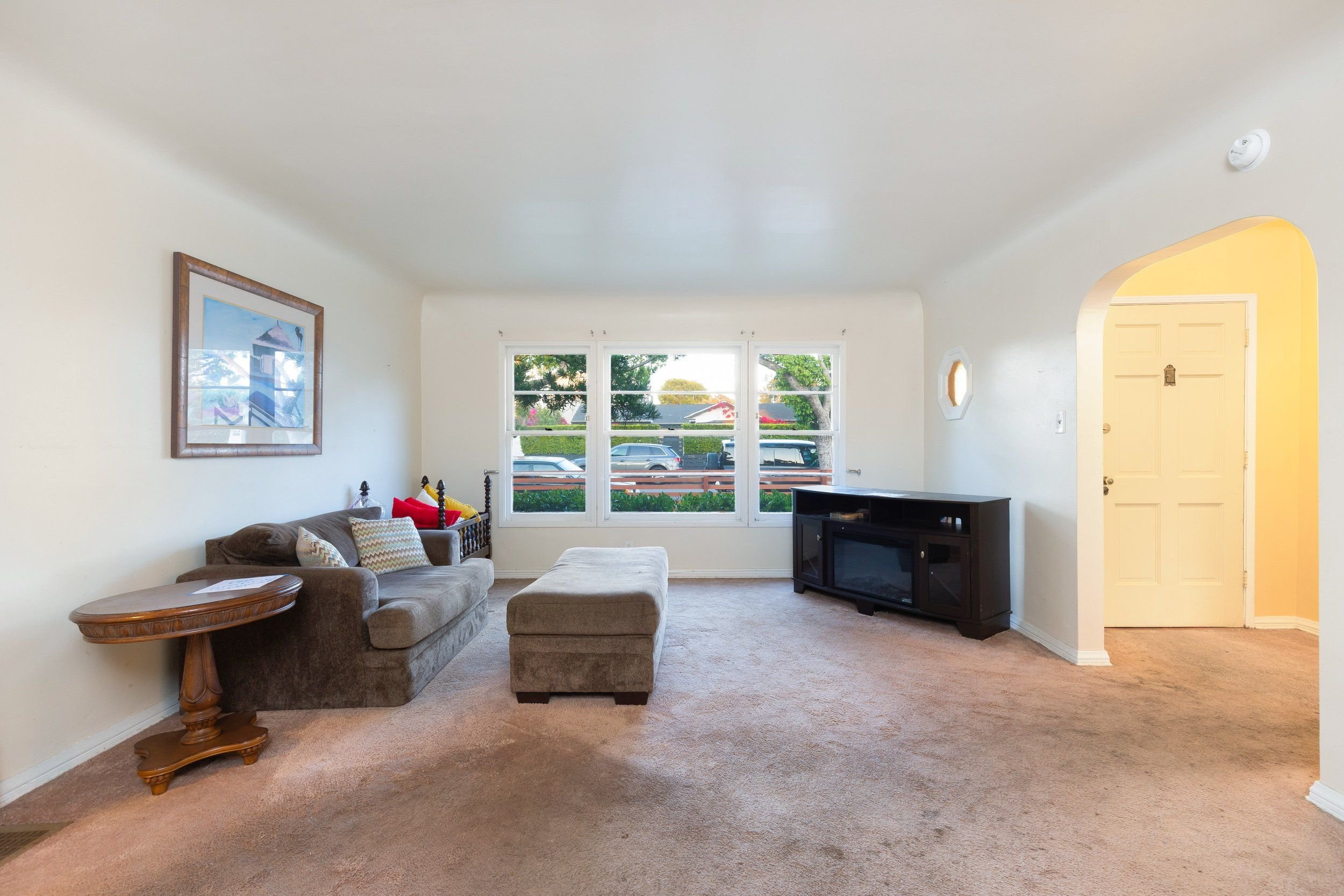 Living room with beige carpet, beige walls, a gray couch with striped pillows, a beige ottoman, a wooden side table, a black TV stand with TV, and multiple windows showing trees and parked cars outside.