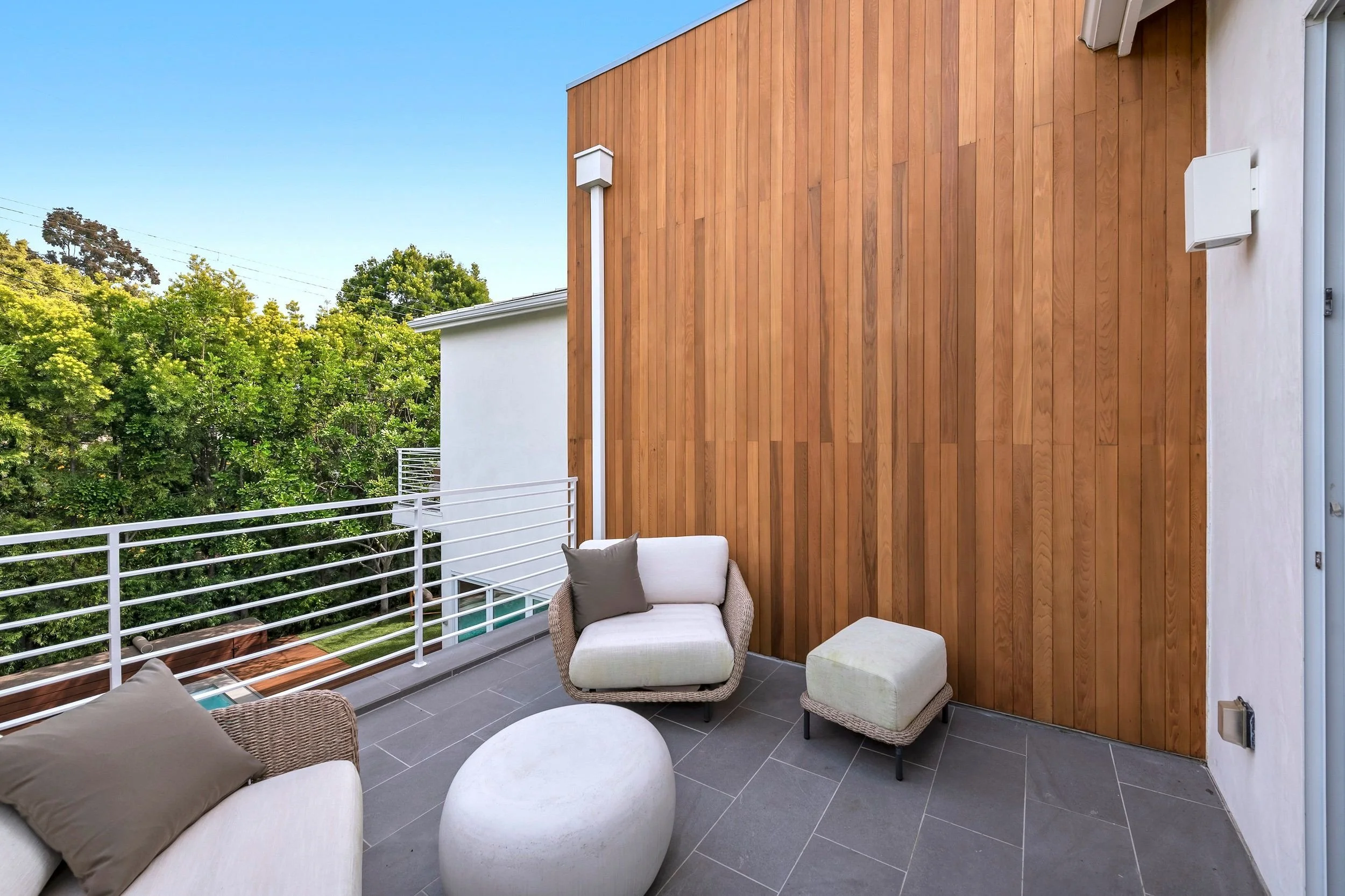Outdoor balcony with modern wicker furniture, cushions, and a small table, surrounded by a white metal railing and a wooden accent wall, with trees and a clear blue sky in the background.
