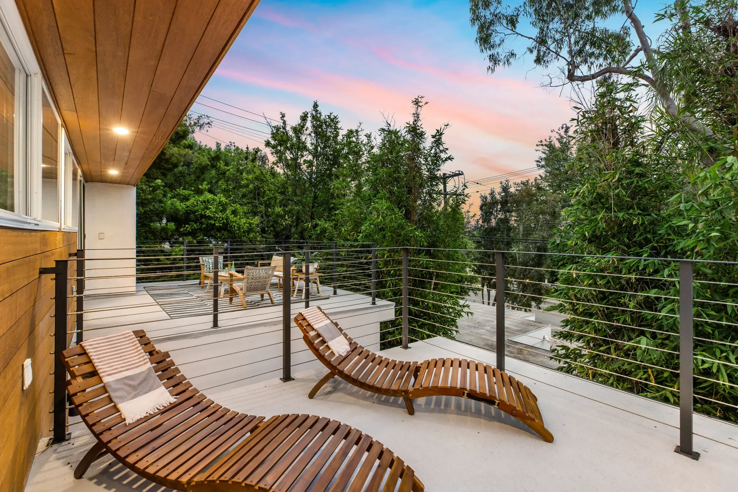Modern balcony with four wooden lounge chairs with towels, surrounded by metal railing, overlooking lush trees, during pink and blue sunset sky.