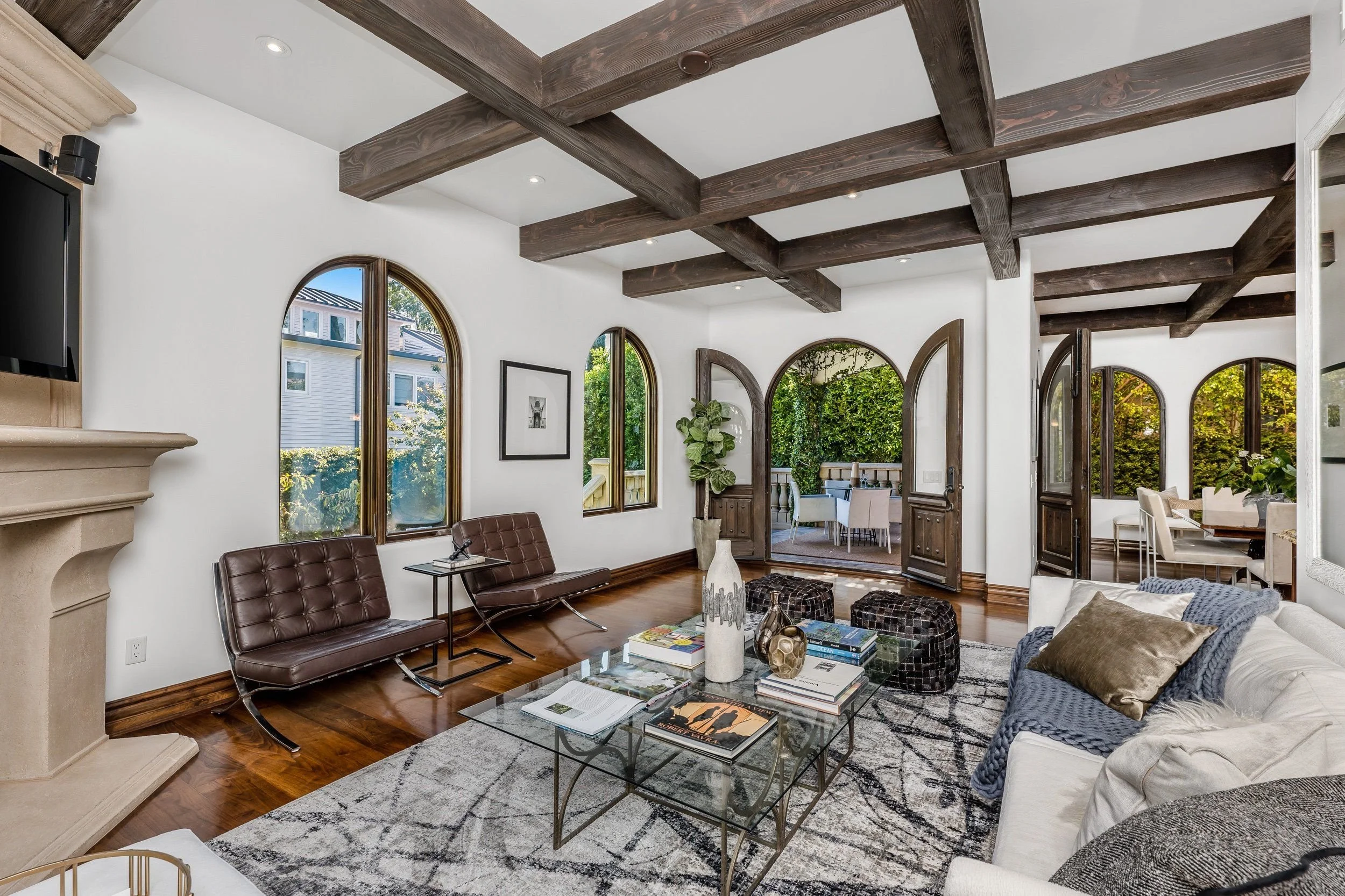 Living room with white walls, dark wooden beam ceiling, arched windows, and furniture including a leather sofa, glass coffee table, and a patterned area rug.