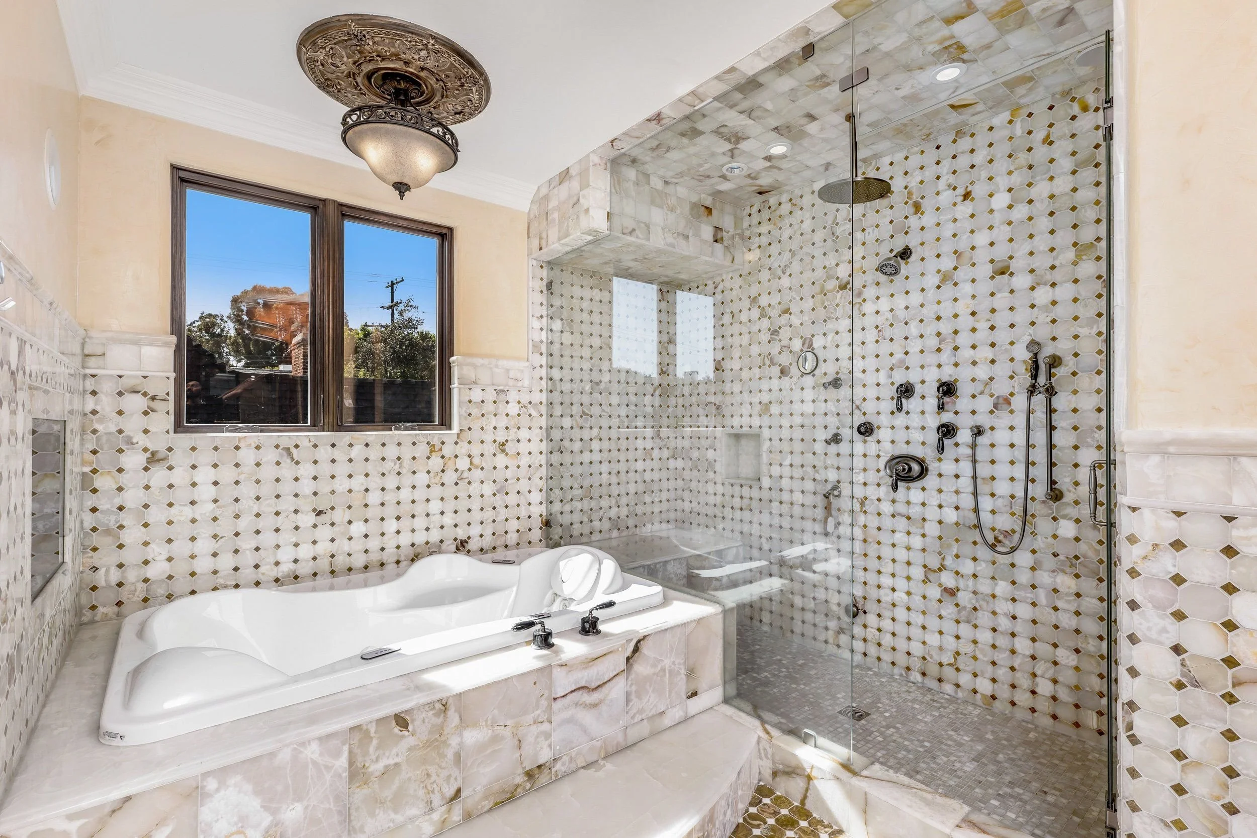 A luxurious bathroom featuring a built-in bathtub, large window, ornate ceiling fixture, and a separate glass-enclosed shower with patterned tile walls.