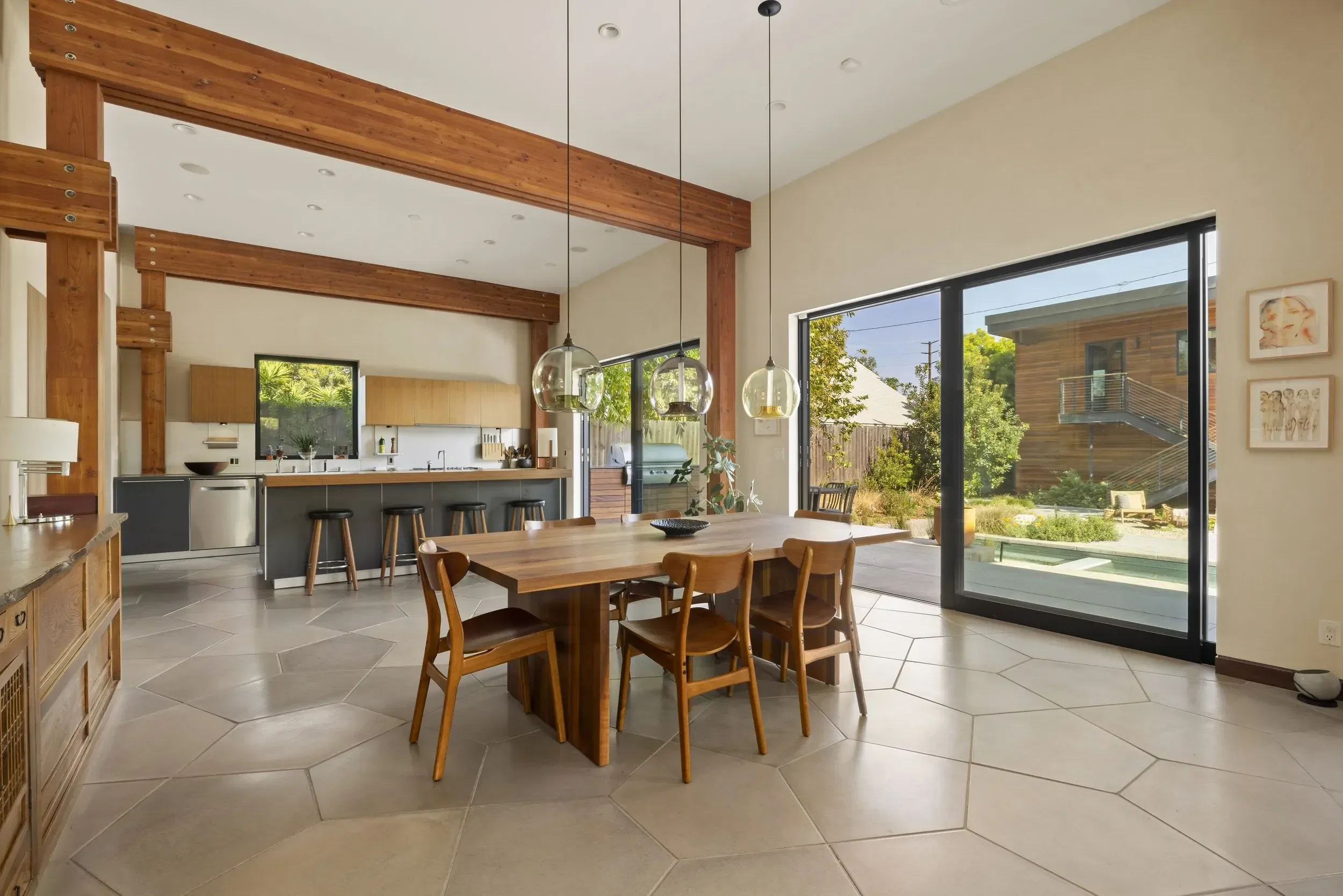Open-concept living and dining area with wooden beams, large windows with a view of a backyard with trees and a patio, and modern furniture including a dining table with chairs.