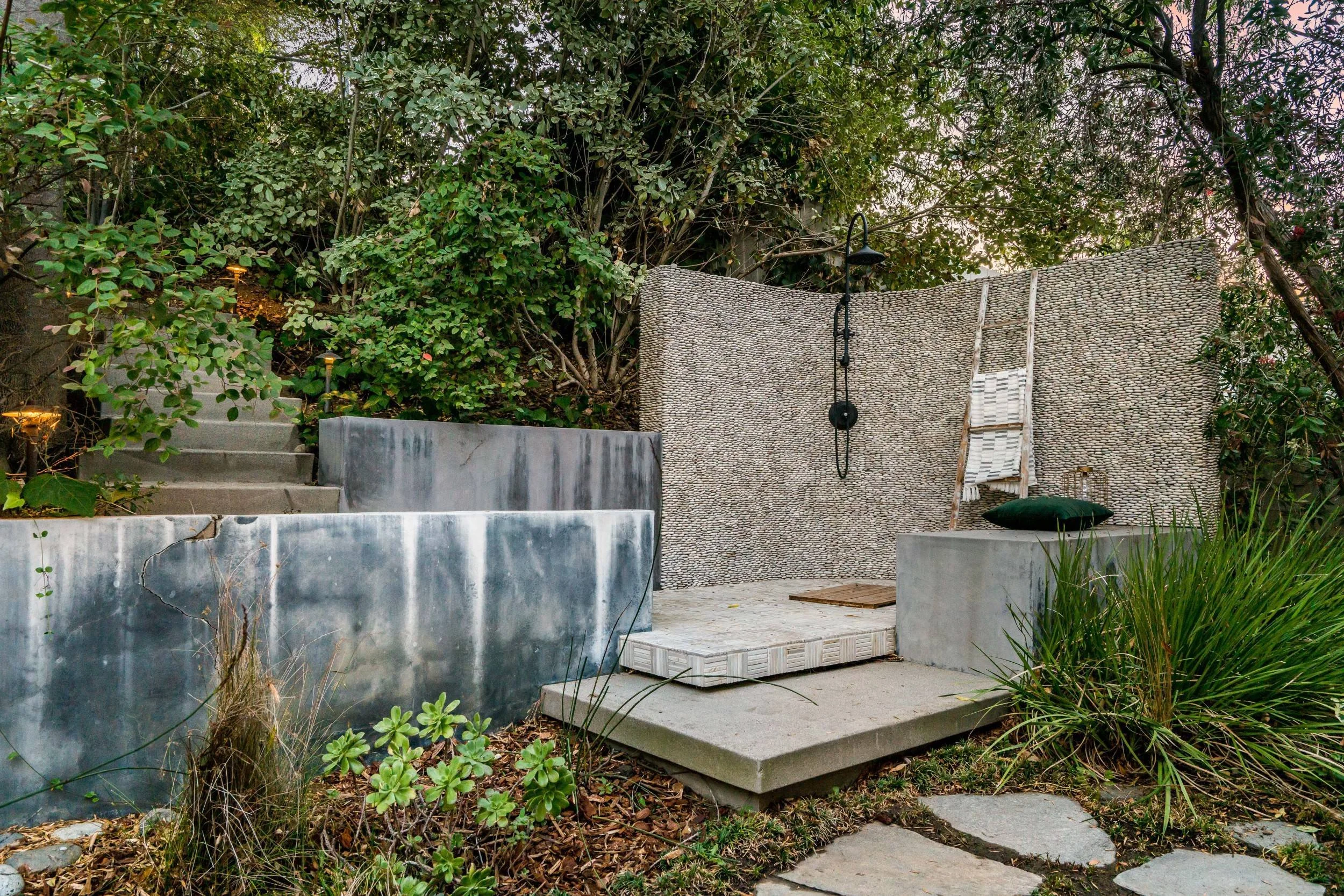 Outdoor patio area with steps, a curved stone wall, a hanging outdoor shower, a green pillow, and surrounding plants and trees.