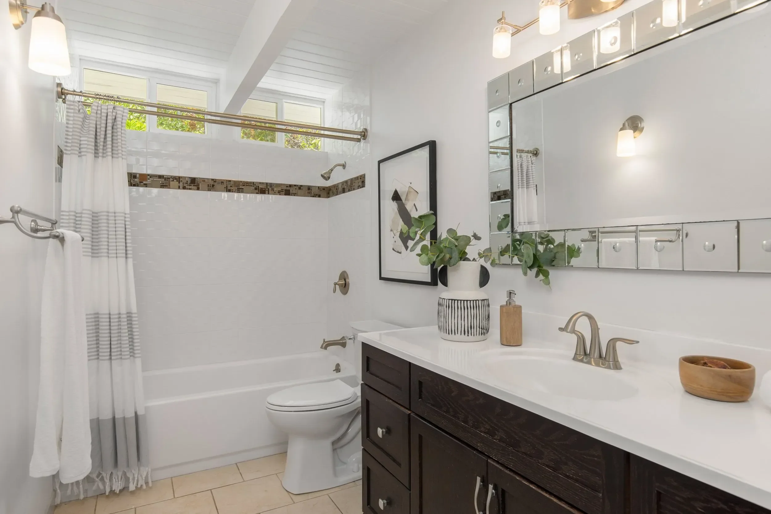 A modern bathroom with white tiled walls and a dark wooden vanity with a white countertop and a large mirror above. There are two wall-mounted lights on either side of the mirror. The bathroom has a tub with a shower curtain and window above, providi
